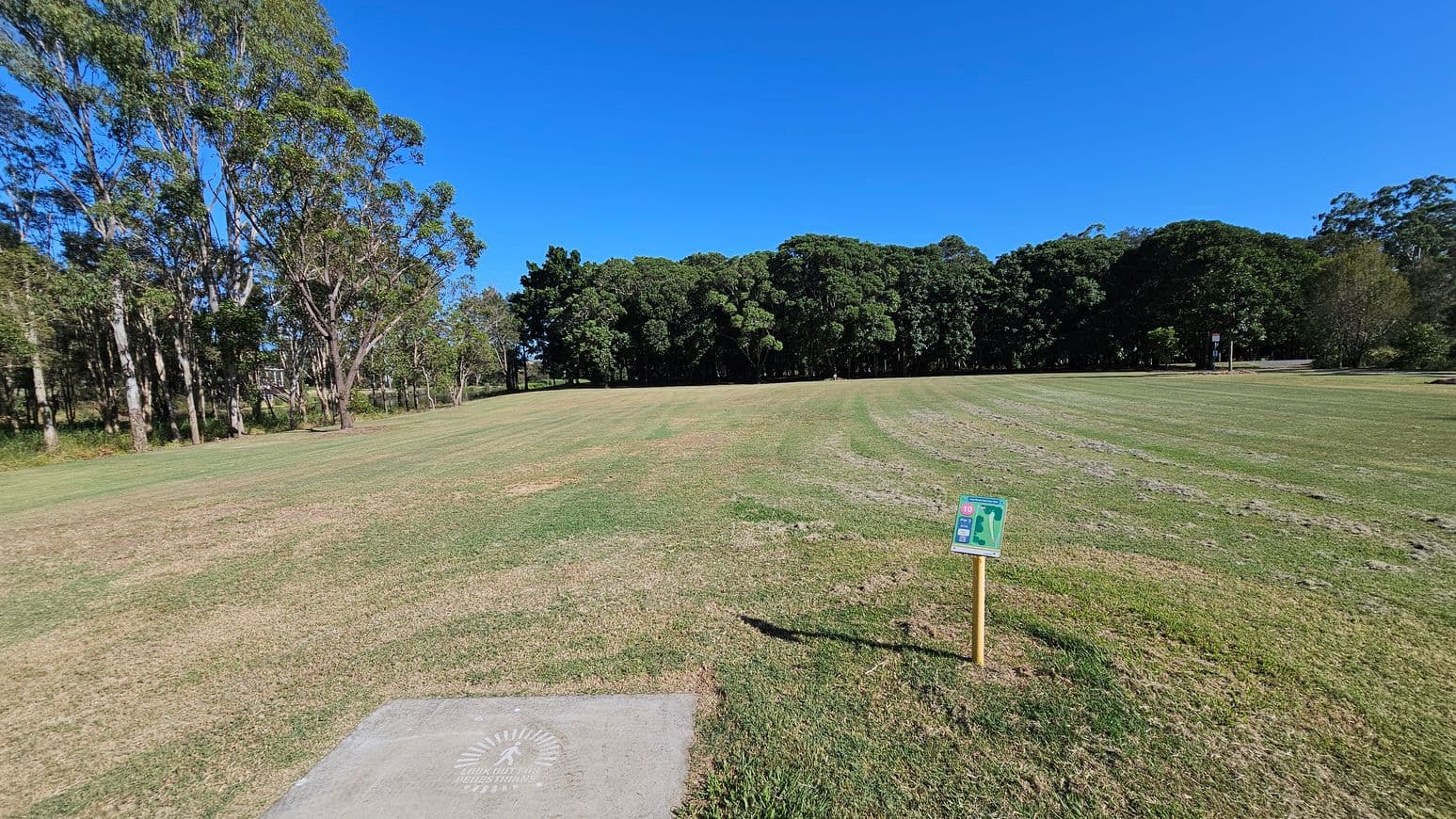 A grassy field with a mix of green and brown patches. On the left, there are tall trees with slender trunks. In the centre, a dense line of trees forms a natural boundary. On the right, a small sign with a map is mounted on a wooden post. The foreground has a concrete slab with a logo or text. The sky is clear and blue.