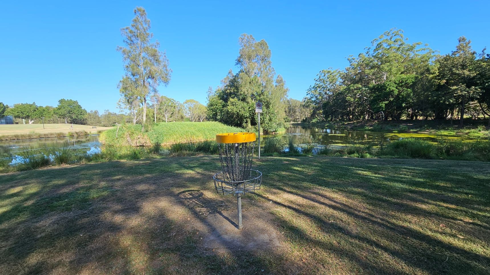 A disc golf basket with a yellow top is in the foreground, standing on a grassy area. In the background, there is a small body of water surrounded by lush greenery and tall trees. Shadows of trees stretch across the grass, and a signpost is visible near the water's edge. The scene is well-lit, suggesting a sunny day.