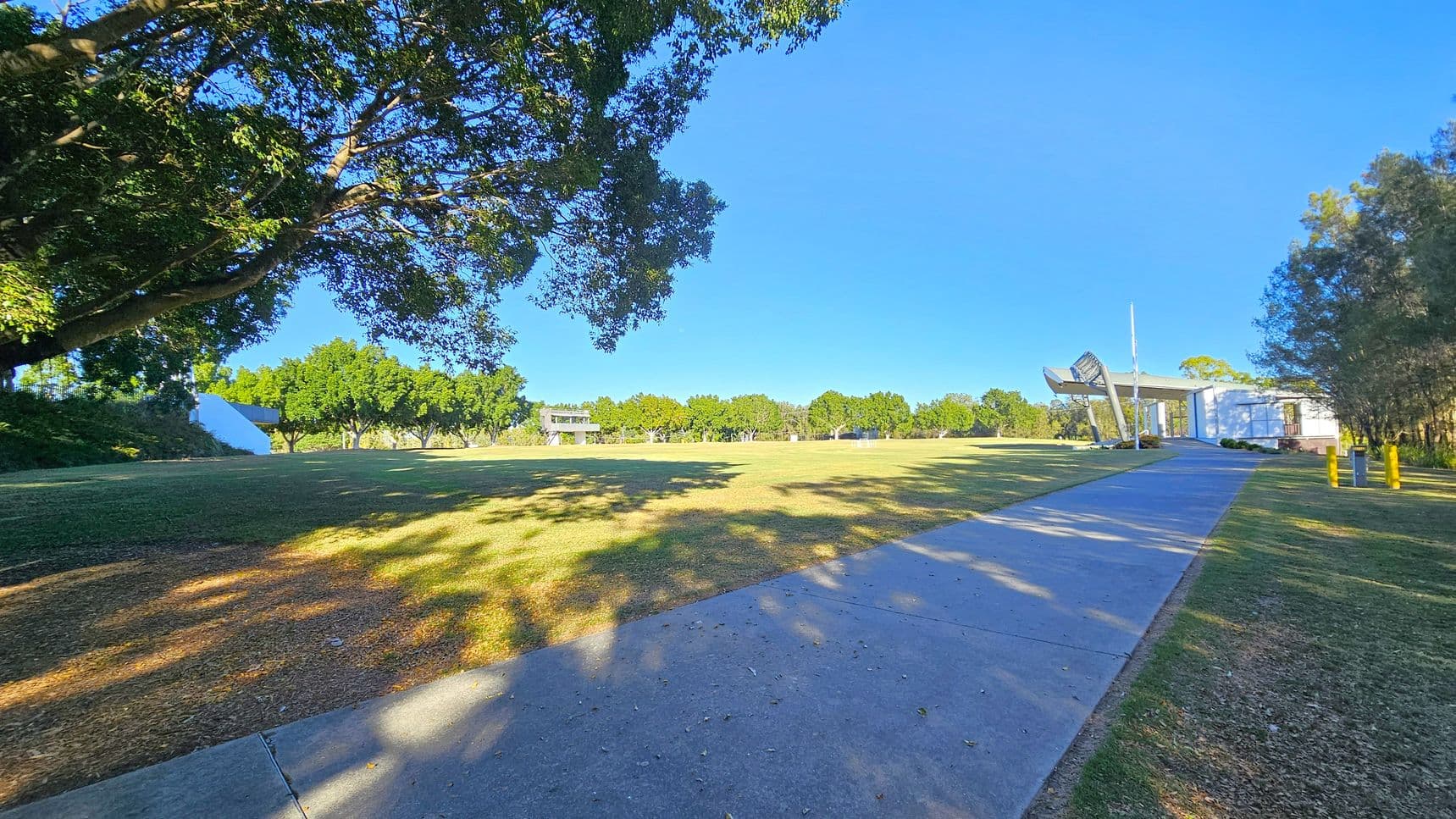 A wide grassy field with scattered trees in the background. On the left, a large tree provides shade over the grass. A concrete path runs from the foreground to the right, leading to a modern structure with a metal sculpture near its entrance. The building is white with a glass facade. The sky is clear and blue, casting shadows on the ground.
