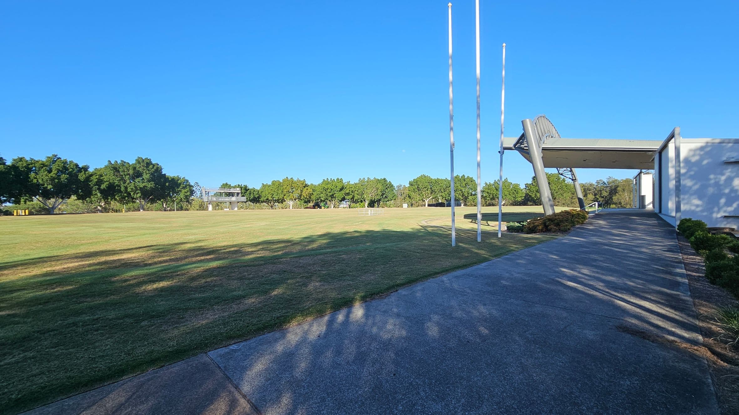 A large grassy field stretches across the image, bordered by a row of trees in the background. On the right, a modern structure with a slanted roof and tall flagpoles stands next to a concrete pathway. The pathway is lined with small shrubs and leads towards the building. Shadows from the trees and structure are cast on the grass and path. The sky is clear and blue.