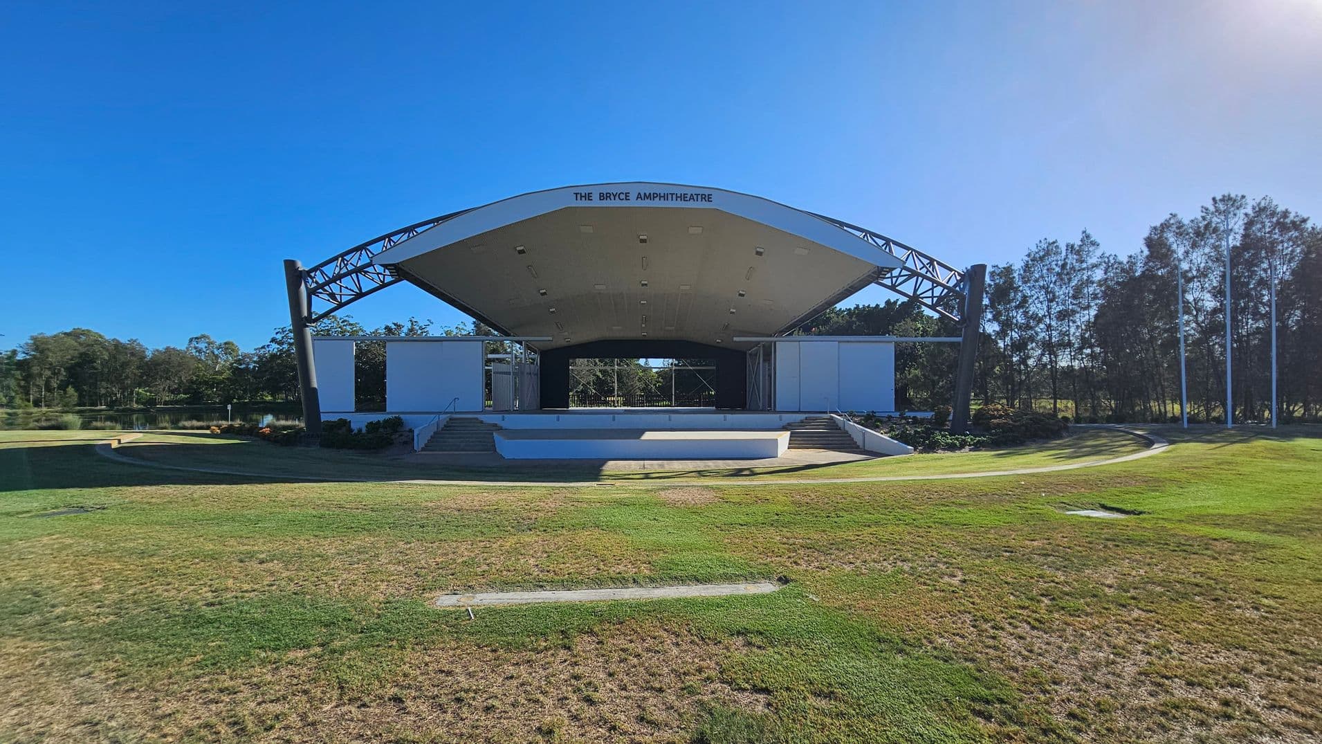 An outdoor amphitheatre with a large, curved roof structure and open sides. The stage is elevated with steps leading up from the grassy foreground. The amphitheatre is surrounded by trees on both sides, and there is a small body of water visible to the left. The grass in the foreground is patchy, with areas of green and brown. The sky is clear and blue.