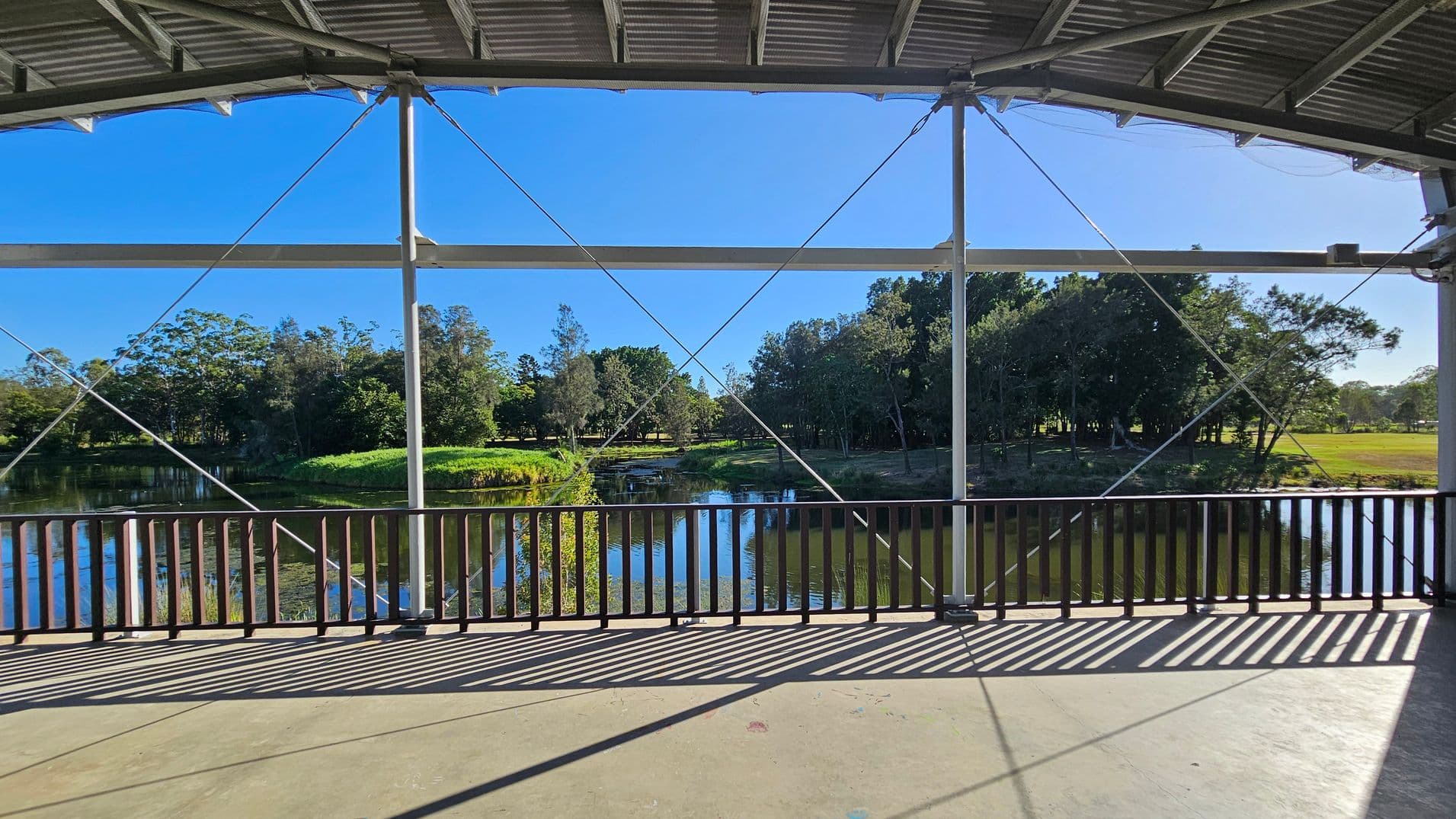 A view from under a metal roof with cross-bracing cables, looking out over a body of water surrounded by lush greenery and trees. The foreground features a concrete floor with shadows cast by a wooden railing. The background shows a clear blue sky and a variety of trees lining the water's edge, creating a serene natural setting.