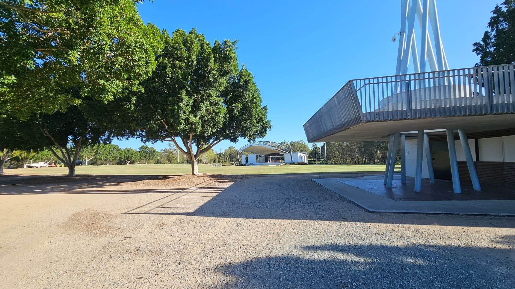 On the left, there are large, leafy trees casting shadows on a gravel path. In the centre, a grassy field extends towards a structure with a curved roof, possibly a stage or pavilion. On the right, a modern structure with a circular platform and metal railings is supported by angled pillars. The ground is a mix of gravel and grass, and the area is surrounded by more trees in the background.