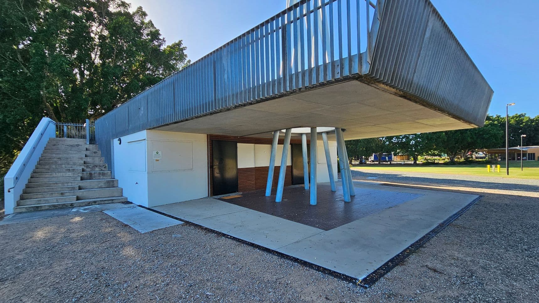 A modern structure with a flat roof supported by angled metal columns. On the left, a set of concrete stairs with a metal railing leads to the upper level. The building has a combination of white and wooden panelling. The ground is covered with gravel, and there are trees in the background, providing shade. A pathway extends to the right, with a few lamp posts and a grassy area beyond.