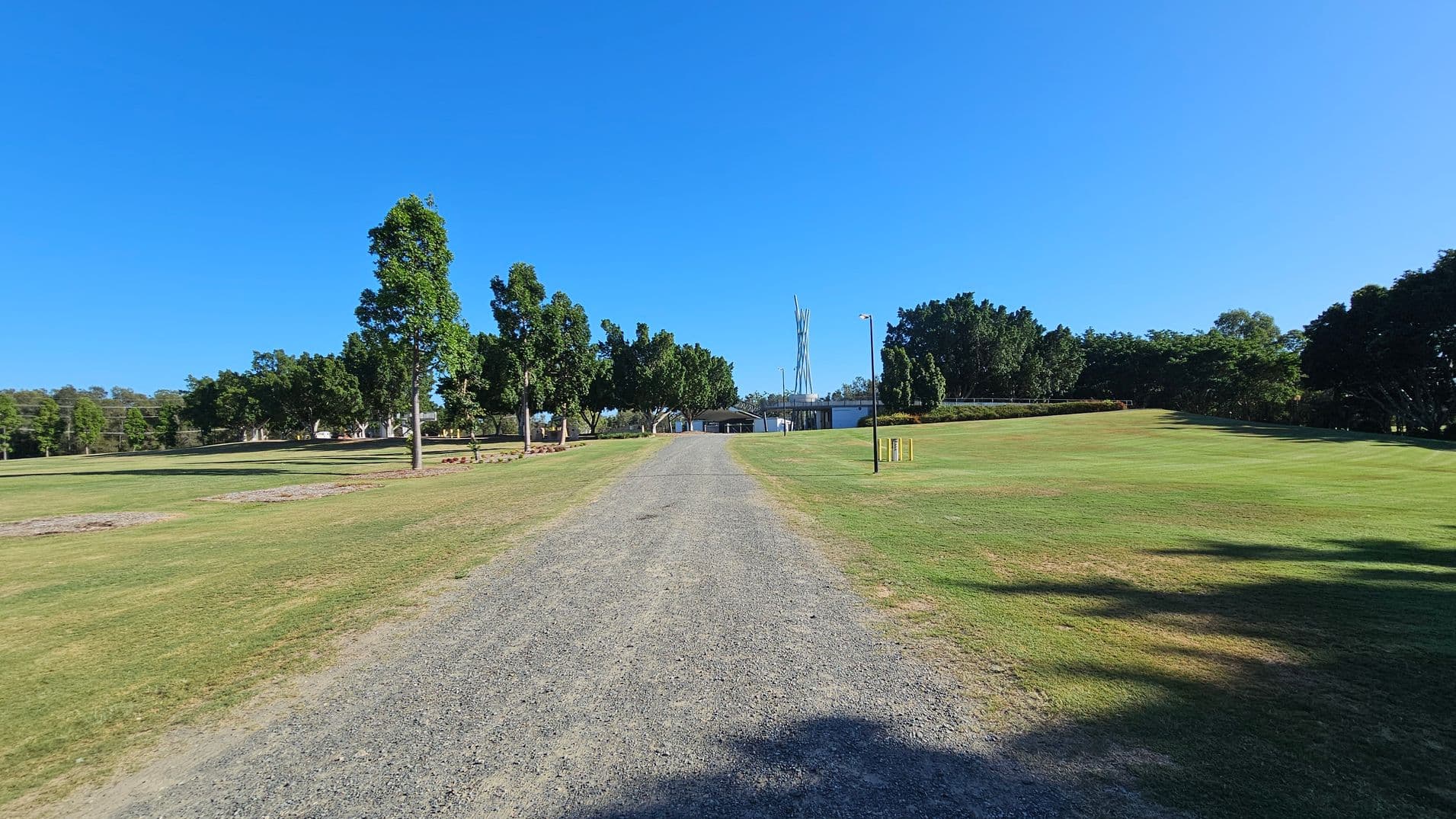 A gravel path runs through the centre of a grassy area, leading towards a structure in the distance. Trees line both sides of the path, with more dense foliage on the right. On the left, the grass appears slightly patchy. In the background, a tall, slender structure is visible, surrounded by more trees and a few buildings. The sky is clear and blue.