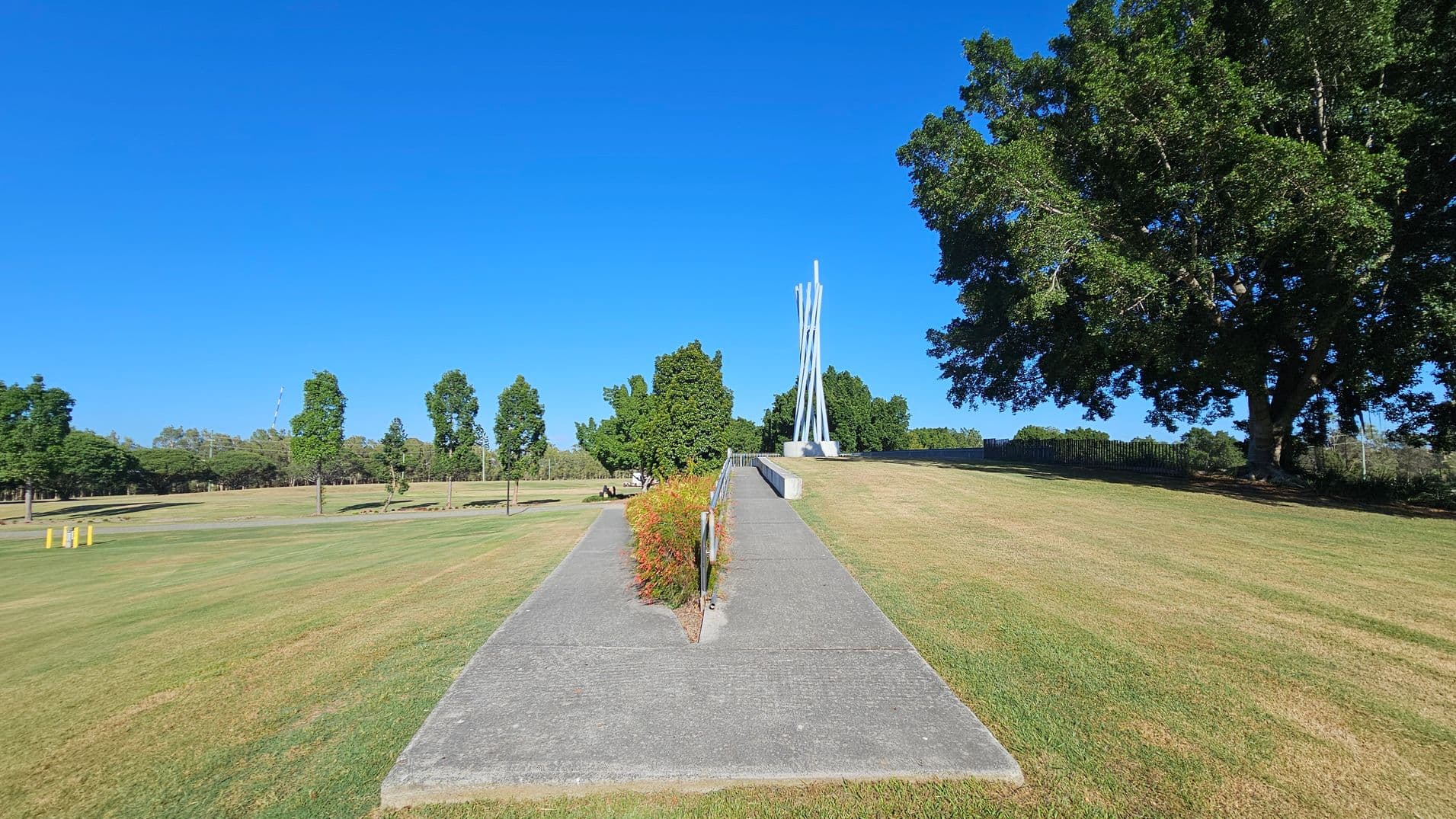 A concrete path leads up a gentle slope, bordered by a metal railing and small shrubs. At the top, a tall, abstract metal sculpture stands prominently. To the right, a large tree with dense foliage provides shade. The left side features a grassy area with scattered trees and a few yellow posts. The background is filled with more trees and a clear blue sky.