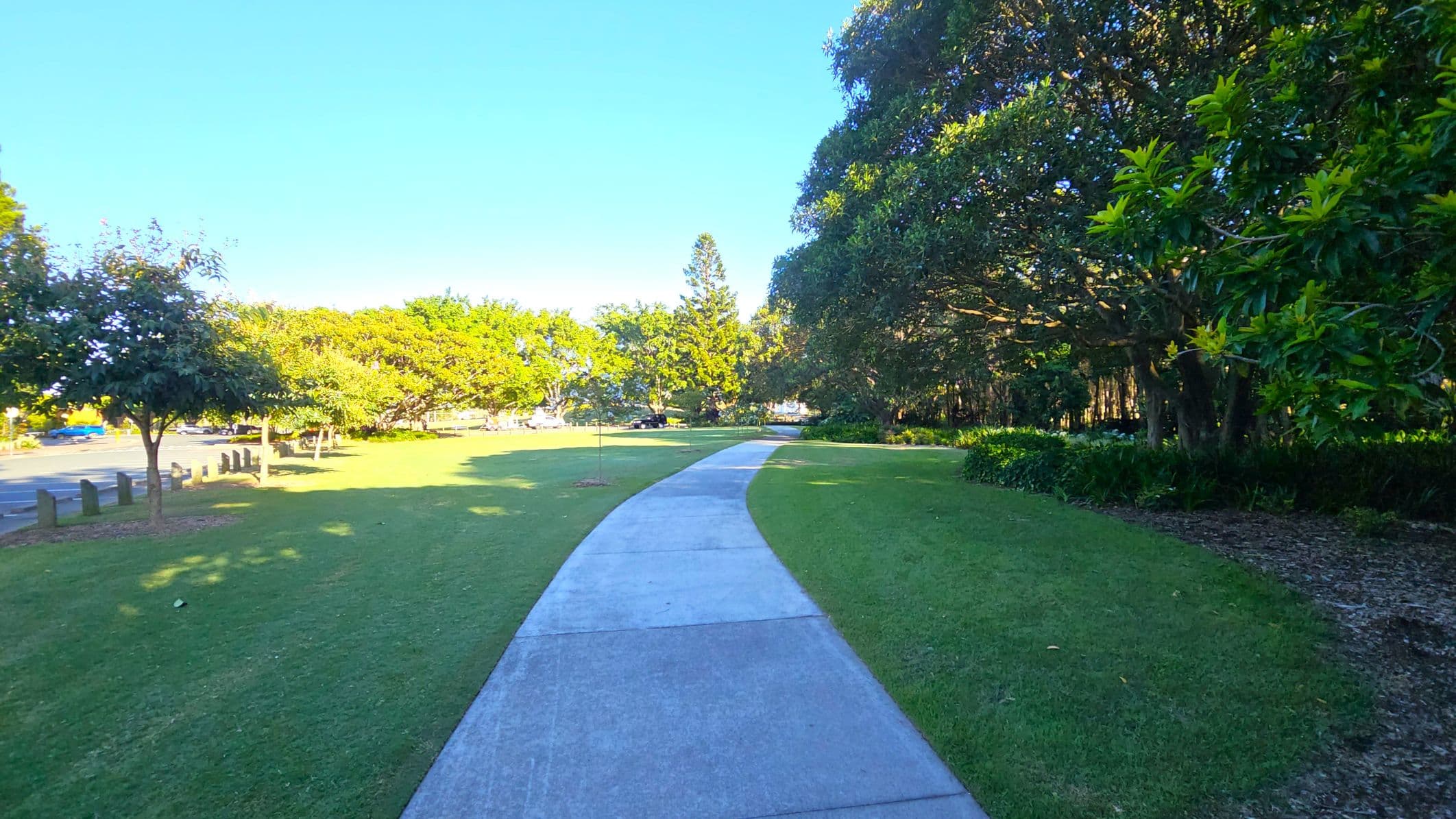 A concrete path curves through a grassy park. On the left, there are small trees and a row of wooden posts near a road with a parked blue car. On the right, dense foliage and larger trees provide shade. The background features more trees and open green space. The scene is well-lit, suggesting a sunny day.