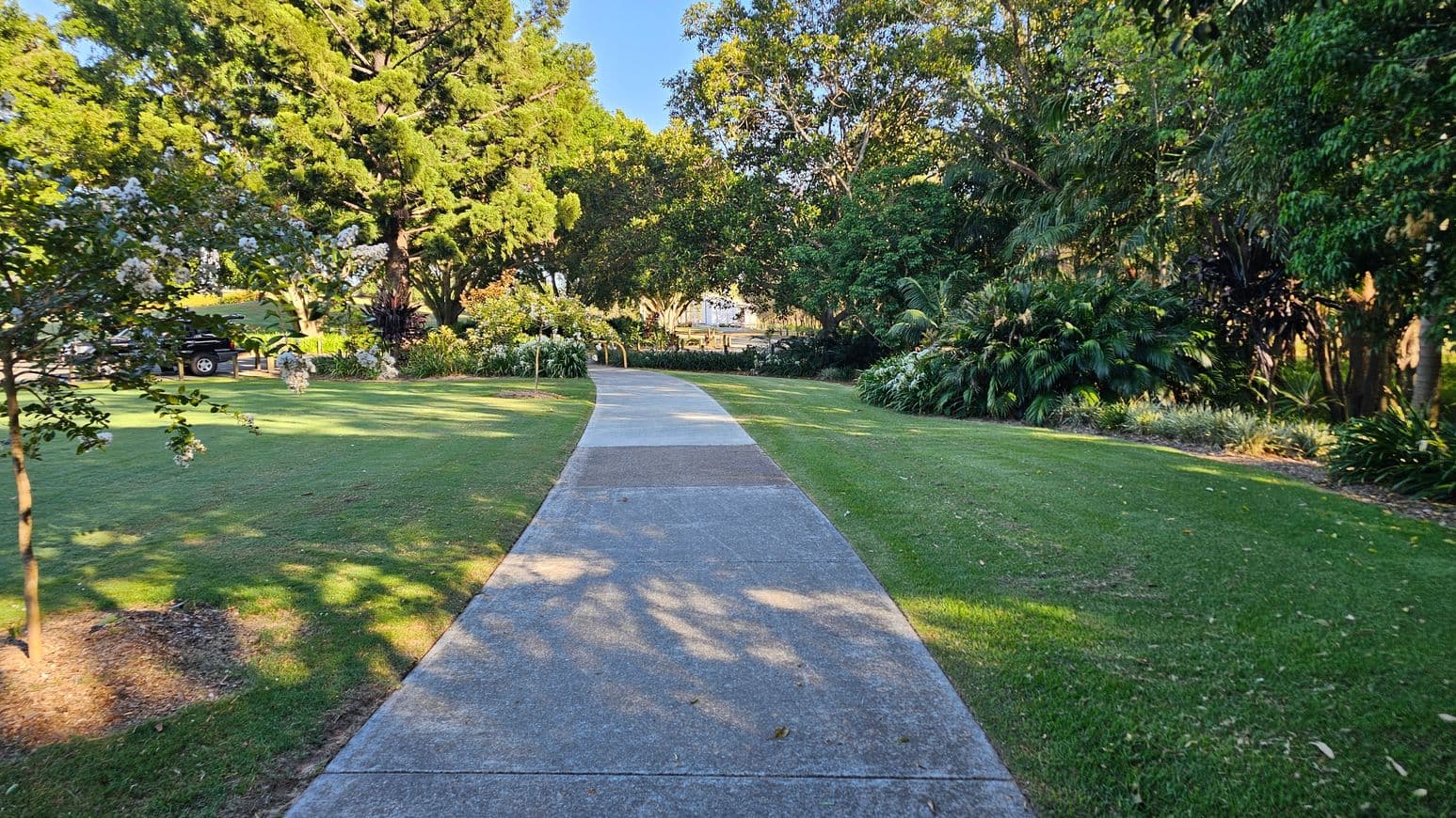 A concrete path runs through a park, flanked by well-maintained grass. On the left, there are flowering trees and a parked vehicle partially visible. On the right, dense foliage and various plants create a lush border. Tall trees with thick canopies provide shade, and sunlight filters through, casting dappled shadows on the path and grass.