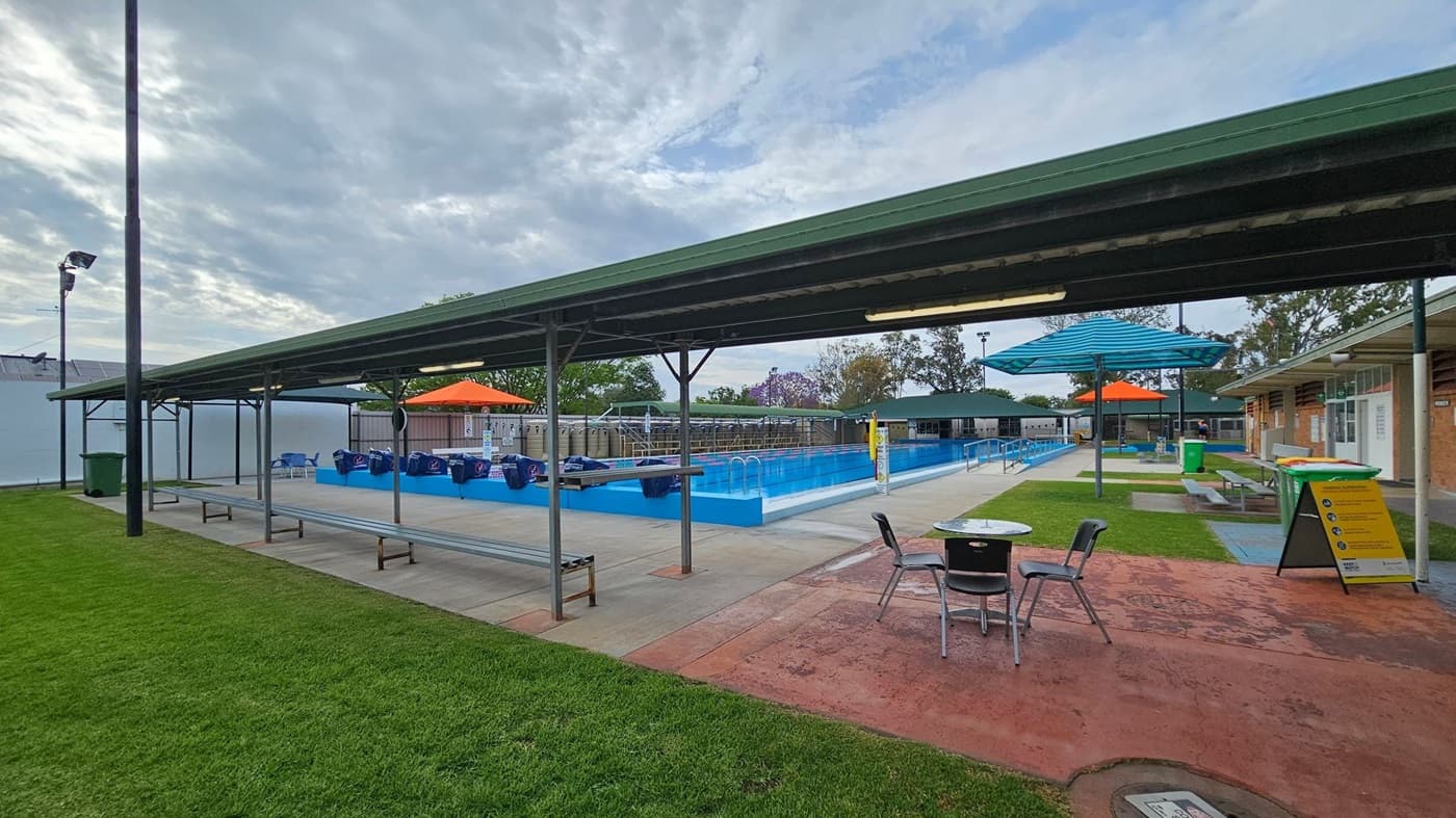 Outdoor swimming pool area with a long, rectangular pool in the centre. On the left, there is a covered seating area with chairs. The right side features a grassy area with a bench and green bins. Blue and orange shade structures are positioned over the poolside. A yellow sign with safety instructions is in the foreground. The background includes trees and a fence, with a cloudy sky above. The ground is a mix of concrete and grass.