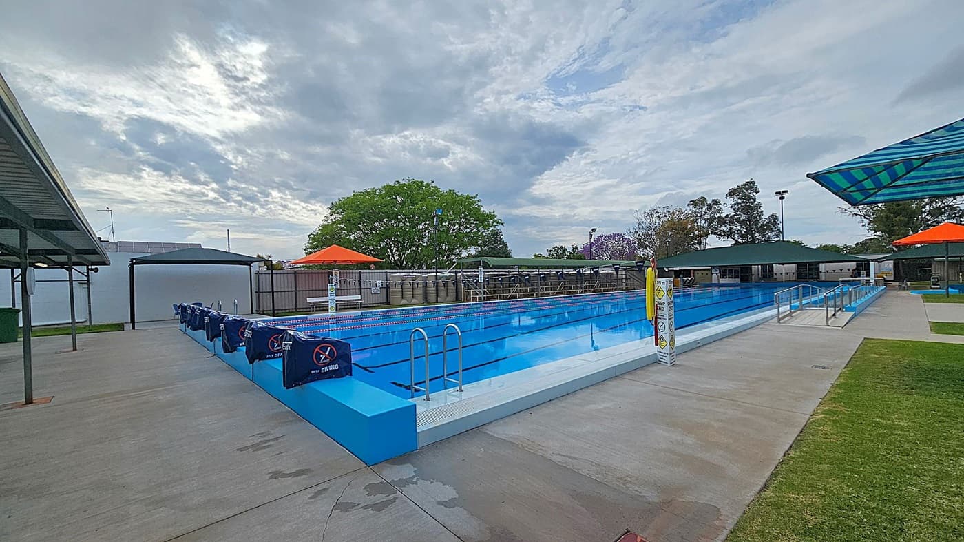 Outdoor swimming pool with clear blue water, surrounded by a concrete deck. On the left, a covered area with a metal roof and a green bin. In the centre, the pool has lane dividers and a "No Diving" sign. On the right, a grassy area and a shaded seating area with a striped canopy. Orange umbrellas and trees are visible in the background.