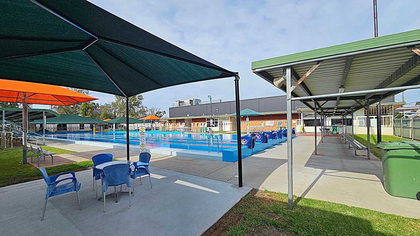 A swimming pool area with blue and white lane dividers in the centre. On the left, there are blue chairs and a table under a large green umbrella, with an orange umbrella nearby. On the right, a covered walkway with a green roof and benches underneath. The background features a brick building with a green roof and a dark grey structure behind it. The ground is a mix of concrete and grass.