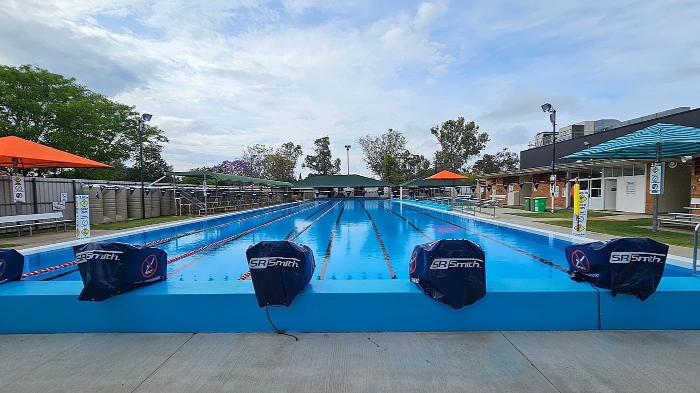 A long, rectangular outdoor swimming pool with clear blue water is in the centre. The pool is divided into lanes by red and white ropes. On the left and right, there are orange and blue shade structures, benches, and signs indicating a depth of 2.5 metres. The background features trees and a building with a dark roof. The pool deck is concrete, and there are covered starting blocks labelled "SR Smith" at the pool's edge.
