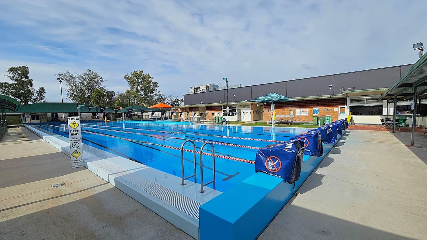 Outdoor swimming pool with clear blue water and lane dividers. A sign indicates a depth of 2.85 metres at the deep end. On the left, a covered seating area with green roofs. On the right, a row of blue covers with no diving signs. In the background, a brick building with a green roof and several trees. The pool deck is concrete.