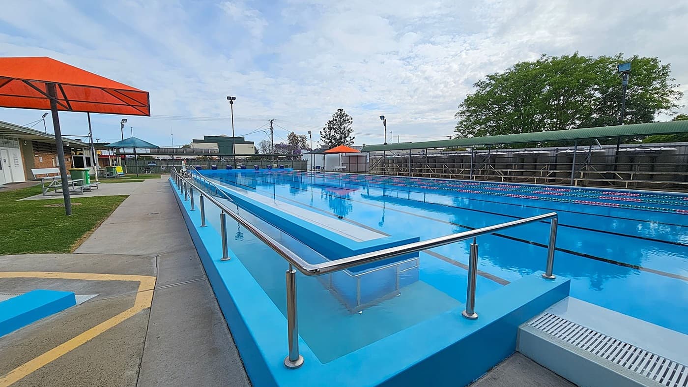 A swimming pool with clear blue water is on the right, featuring a ramp with metal railings. On the left, there is a concrete path and grassy area with orange and blue shade structures. Benches and a small building are visible in the background. A large tree and some fencing are in the distance, along with some outdoor lights.