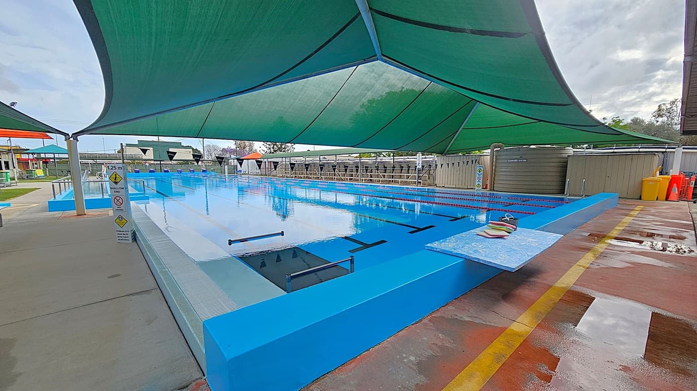 An outdoor swimming pool with clear blue water is covered by large green shade sails. The pool is surrounded by a wet concrete deck. On the right, there are colourful cleaning tools on a blue diving board. Signs indicate a shallow end and safety warnings. In the background, there are seating areas and a fence. The pool lanes are marked with black lines, and the area is well-maintained.