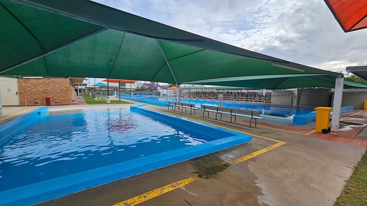 A swimming pool area with blue-tiled pools on the left and right. Green shade sails cover the pools, providing shelter. In the centre, there are benches along the poolside. A yellow bin is visible on the right. The ground is a mix of concrete and grass, with a brick building in the background. The area is well-maintained and organised.