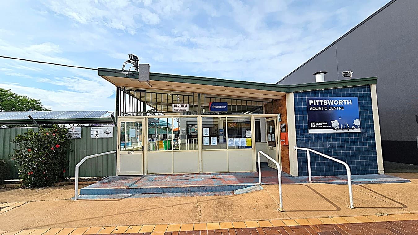Entrance to a building with a sign reading "Pittsworth Aquatic Centre" on the right, featuring blue tiles. The entrance has glass doors with various notices and signs. A ramp with white railings leads up to the doors. On the left, there is a fence with solar panels and a bush with red flowers. The ground is paved with a mix of concrete and tiles. The roof has a green trim and a security camera is mounted above the entrance.
