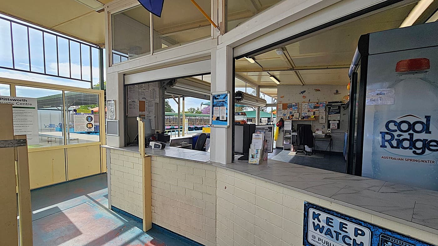 Interior of a reception area at an aquatic centre. On the left, a large window with a sign for "Pittsworth Aquatic Centre" and a view of the pool area outside. In the middle, a counter with a small electronic device and brochures. The right side features a vending machine with a "Cool Ridge" water advertisement. The walls are light-coloured brick, and the ceiling has fluorescent lights. Various notices and posters are displayed on the walls.