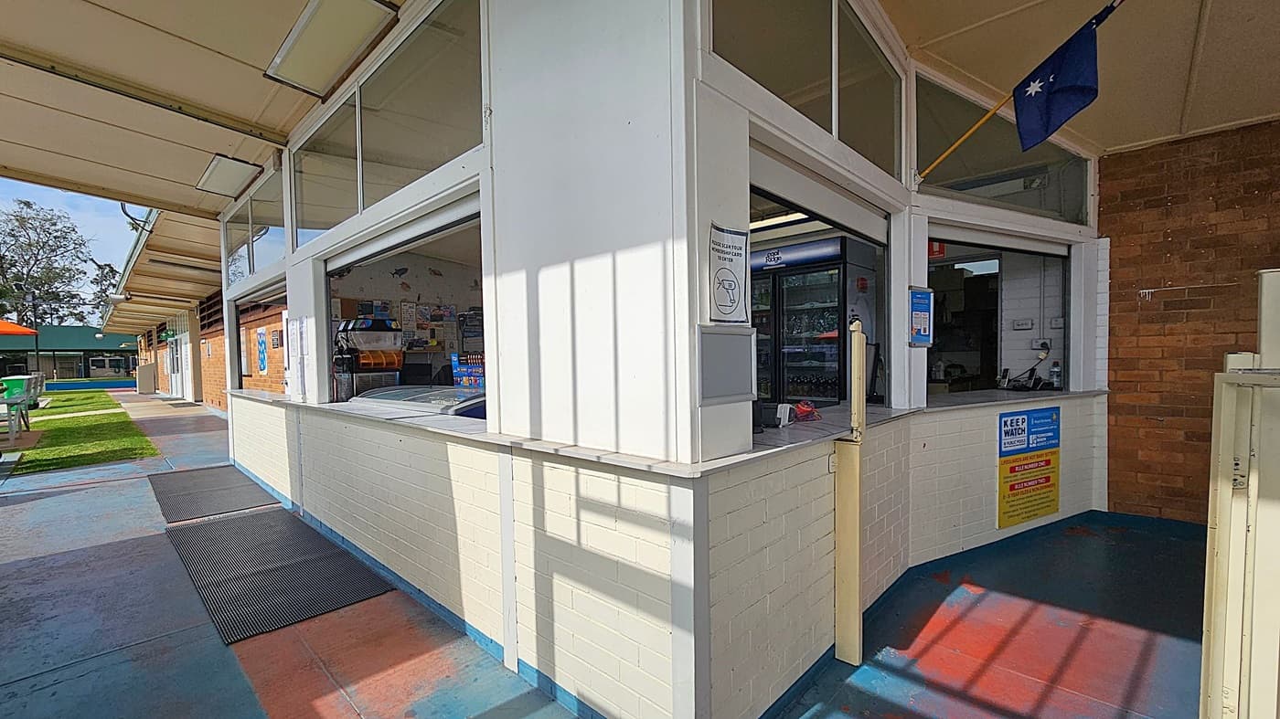 A kiosk with a white and light yellow brick exterior, featuring two open service windows. Inside, a fridge and various items are visible. An Australian flag is displayed above. The floor is a mix of blue and red tiles, with black mats in front of the windows. To the left, there is a walkway with green grass and outdoor seating under orange umbrellas. The ceiling is a light-coloured structure extending over the area.