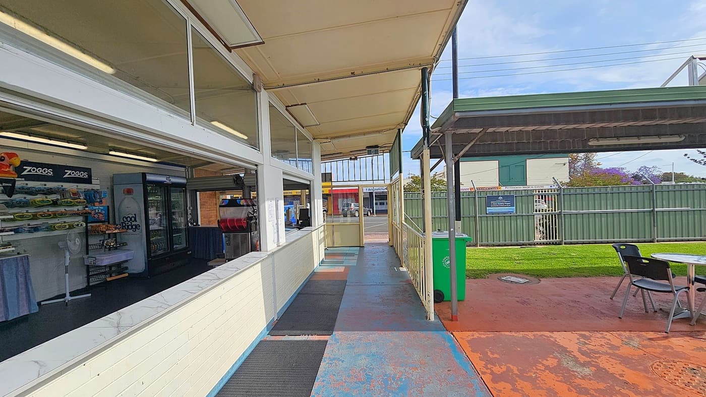 A covered walkway with a shop on the left, displaying shelves of products and a fridge with a "Cool Ridge" label. The floor is painted blue with black mats. On the right, there's a green bin and a seating area with a table and chairs on a reddish-brown concrete surface. A green metal fence and a building are visible in the background. The ceiling is beige with fluorescent lights.