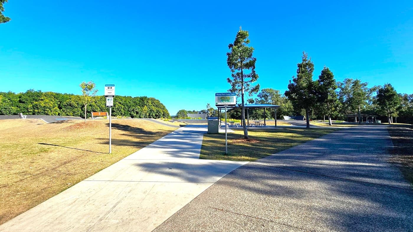 A park scene with a paved path running through the centre. On the left, there's a grassy area with a bench and a signpost. In the middle, a covered seating area with tables is visible. On the right, more trees line another path. The background features a dense line of trees and a clear blue sky. The ground is a mix of grass and concrete paths.
