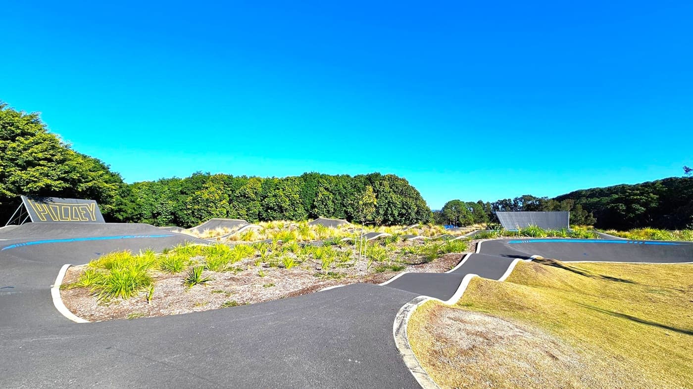 A paved pump track with undulating paths and curves is in the foreground, surrounded by patches of grass and small plants. On the left, a large ramp with the word "PIZZLEY" is visible. In the background, dense green trees form a natural border. The sky is clear and blue.