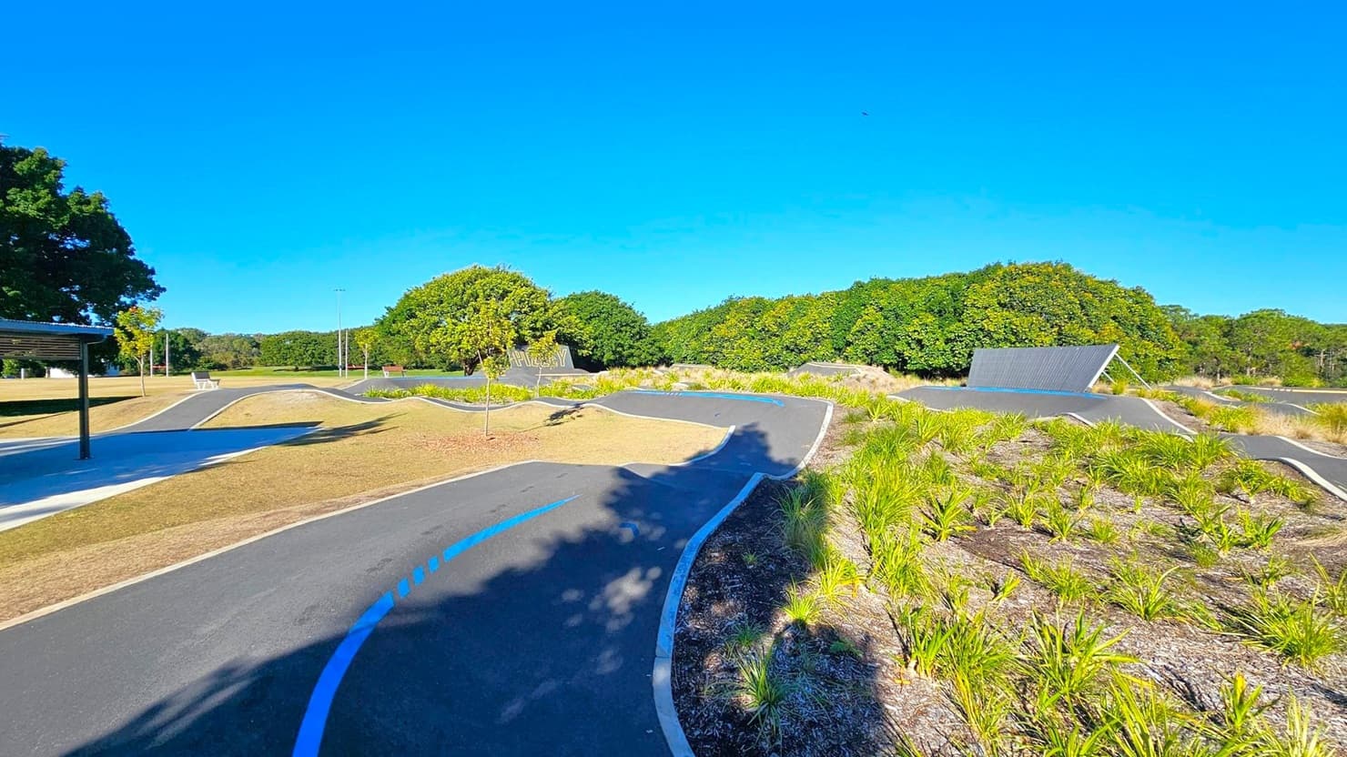 A winding asphalt path with blue markings runs through a landscaped area with grass and small plants. On the left, there's a covered seating area with benches and a few young trees. In the background, dense green trees line the horizon. A large, angled metal structure is visible on the right side of the path. The ground is a mix of grass and mulch, and the sky is clear and blue.