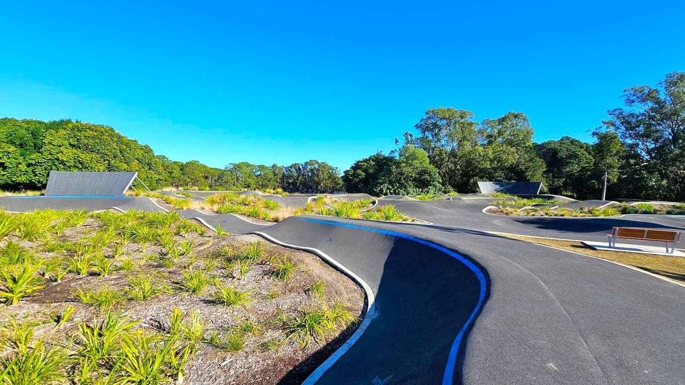 A winding asphalt pump track with smooth curves and undulations runs through the centre, surrounded by patches of grass and small plants. On the left, a sloped structure with a dark roof is visible. To the right, a wooden bench sits on a concrete pad. In the background, dense green trees form a natural border around the area. The sky is clear and blue.
