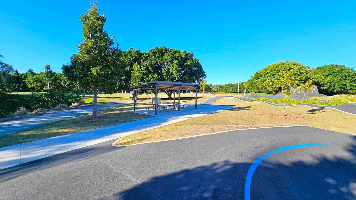 A park scene with a paved path curving through the foreground. On the left, a tree stands beside the path, casting shadows on the ground. In the centre, a sheltered picnic area with tables and benches is visible. The background features a variety of trees and a grassy area. On the right, a sign with text is partially visible, surrounded by more trees and shrubs. The sky is clear and blue.