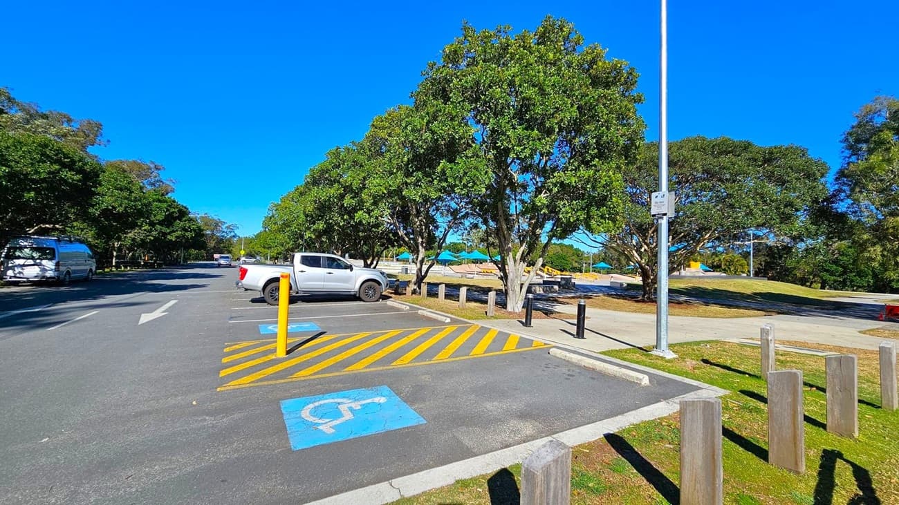 A car park with a designated disabled parking space marked in blue on the asphalt. A white pickup truck is parked nearby. Yellow diagonal lines are painted next to the disabled space. Trees with dense green foliage line the area, providing shade. A tall streetlight stands near the parking area. In the background, a playground with colourful equipment is visible. Wooden posts are placed along the grassy edge of the parking area.