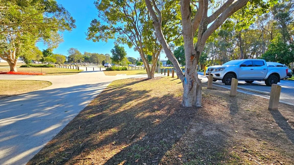 A park scene with a concrete path curving through the centre. On the right, a white pickup truck is parked on the road beside wooden bollards. Trees with green foliage are scattered throughout, casting shadows on the ground. The grass is patchy, with some areas of bare soil. In the background, more trees and open grassy areas are visible under a clear blue sky.