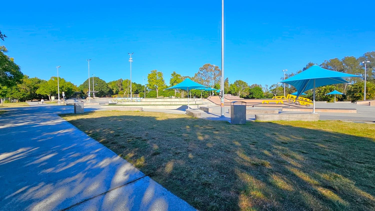 A skate park with concrete paths and ramps is in the centre, featuring blue shade structures. Yellow benches are visible near the ramps. The foreground has a grassy area with shadows from nearby trees. On the left, a paved path runs alongside more grass and trees. In the background, there are tall trees and several light poles.