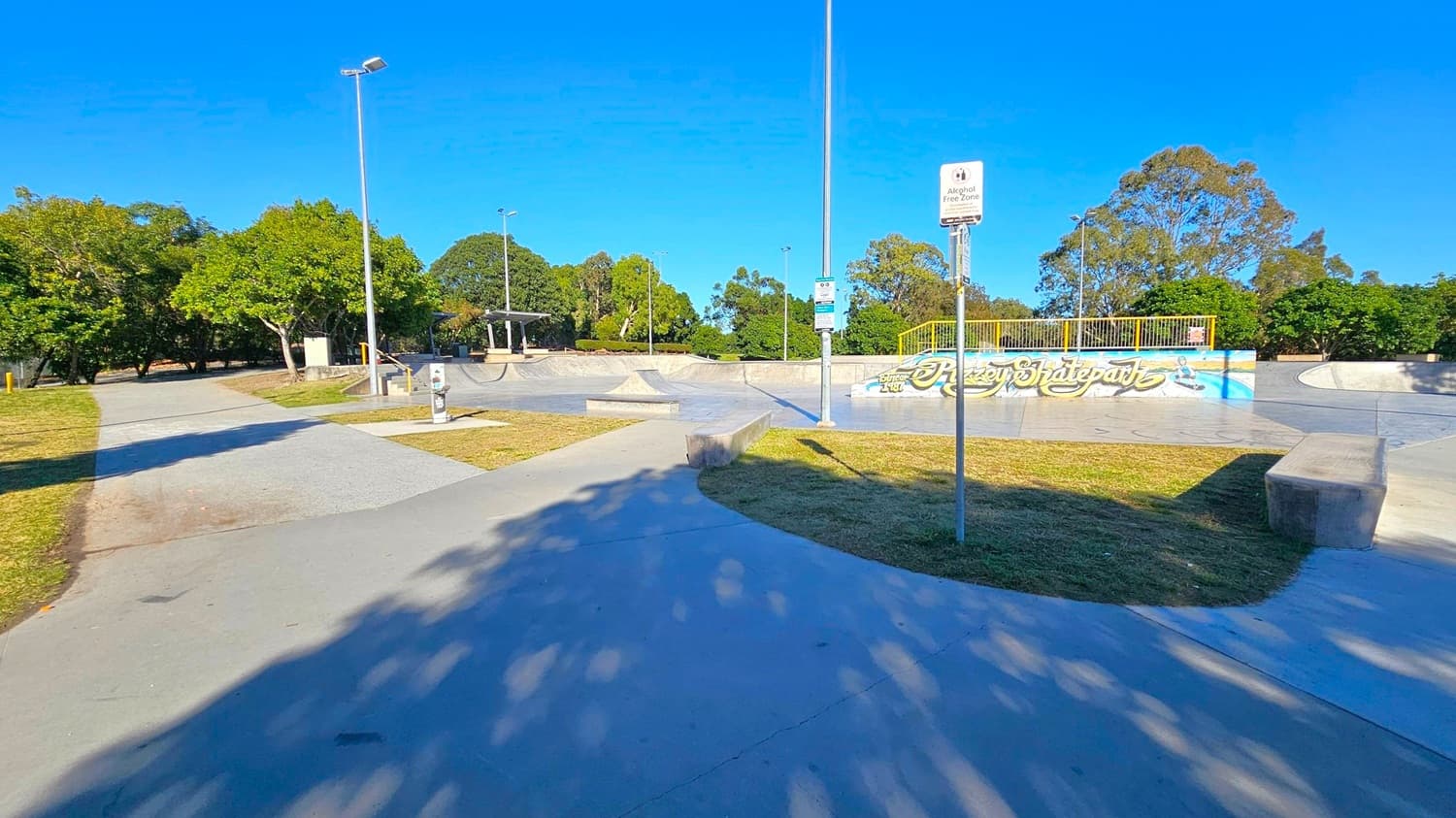 A skatepark with concrete ramps and a graffiti-covered wall in the centre. A signpost with "Alcohol Free Zone" is visible. Trees and greenery surround the area, with a few tall lampposts scattered around. The ground is a mix of concrete paths and grassy patches. Shadows from the trees are cast on the ground.