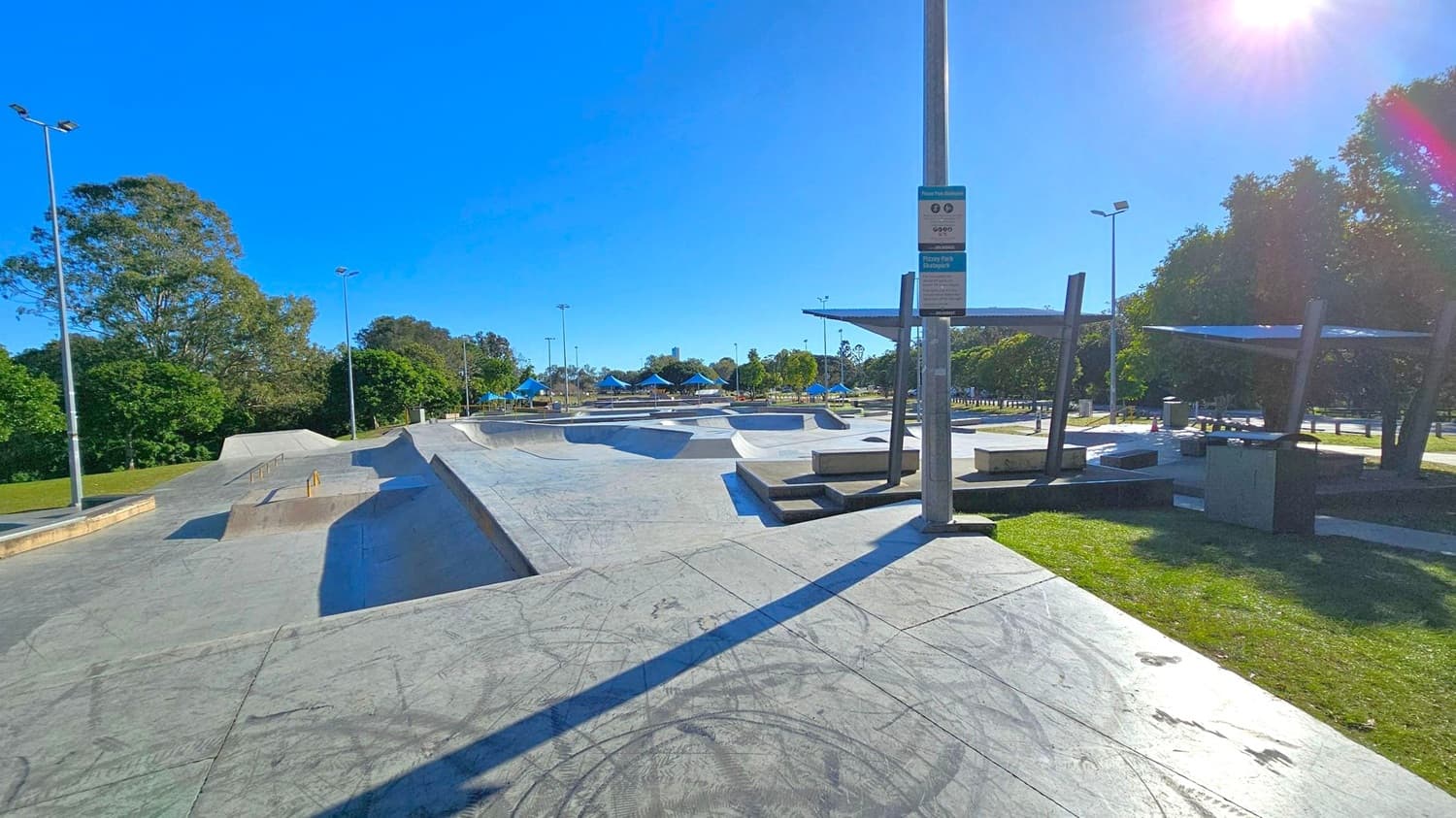 Concrete skate park with ramps and rails in the centre. On the left, there are tall trees and a grassy area. On the right, there are shaded seating areas with metal structures and more trees. A signpost is visible in the foreground. The ground is marked with skate tracks, and there are several tall lampposts throughout the park.
