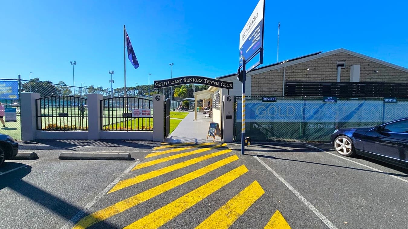 Entrance to the Gold Coast Seniors Tennis Club with a sign above the gate. On the left, a flagpole with a flag and a view of tennis courts behind a fence. On the right, a brick building with a signboard and a parked car. The foreground features a car park with yellow diagonal lines on the asphalt.
