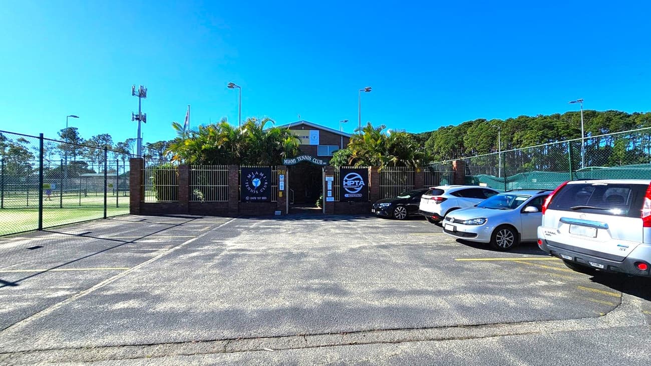 A car park with several vehicles is in the foreground, leading to the entrance of the Miami Tennis Club. The entrance is flanked by palm trees and has signs on either side. A chain-link fence surrounds the tennis courts on the left. In the background, there are tall trees and a clear blue sky.
