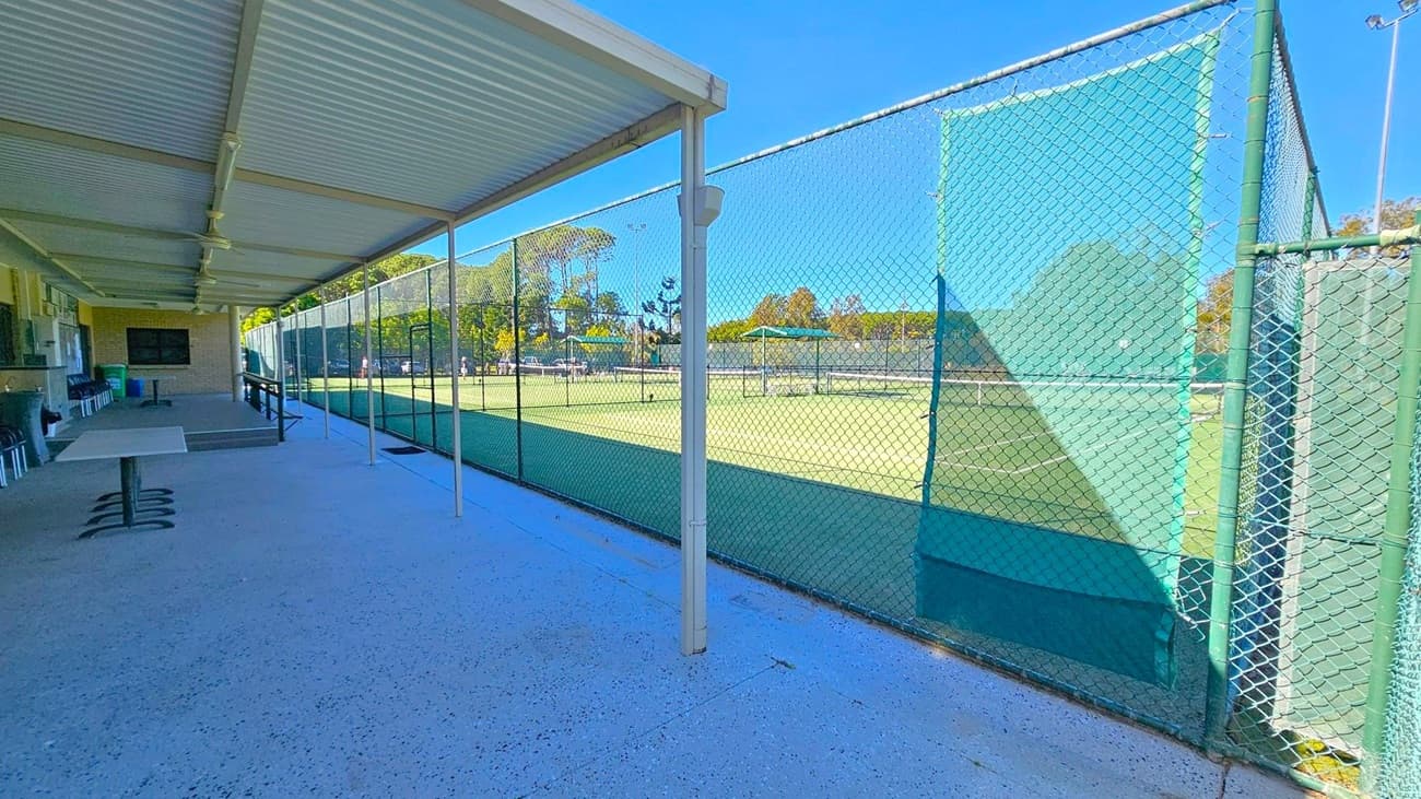 On the left, there is a covered area with a corrugated metal roof, featuring a table and chairs along a brick wall. The floor is concrete. On the right, a chain-link fence with green mesh panels encloses a tennis court. In the background, trees and additional tennis courts are visible.