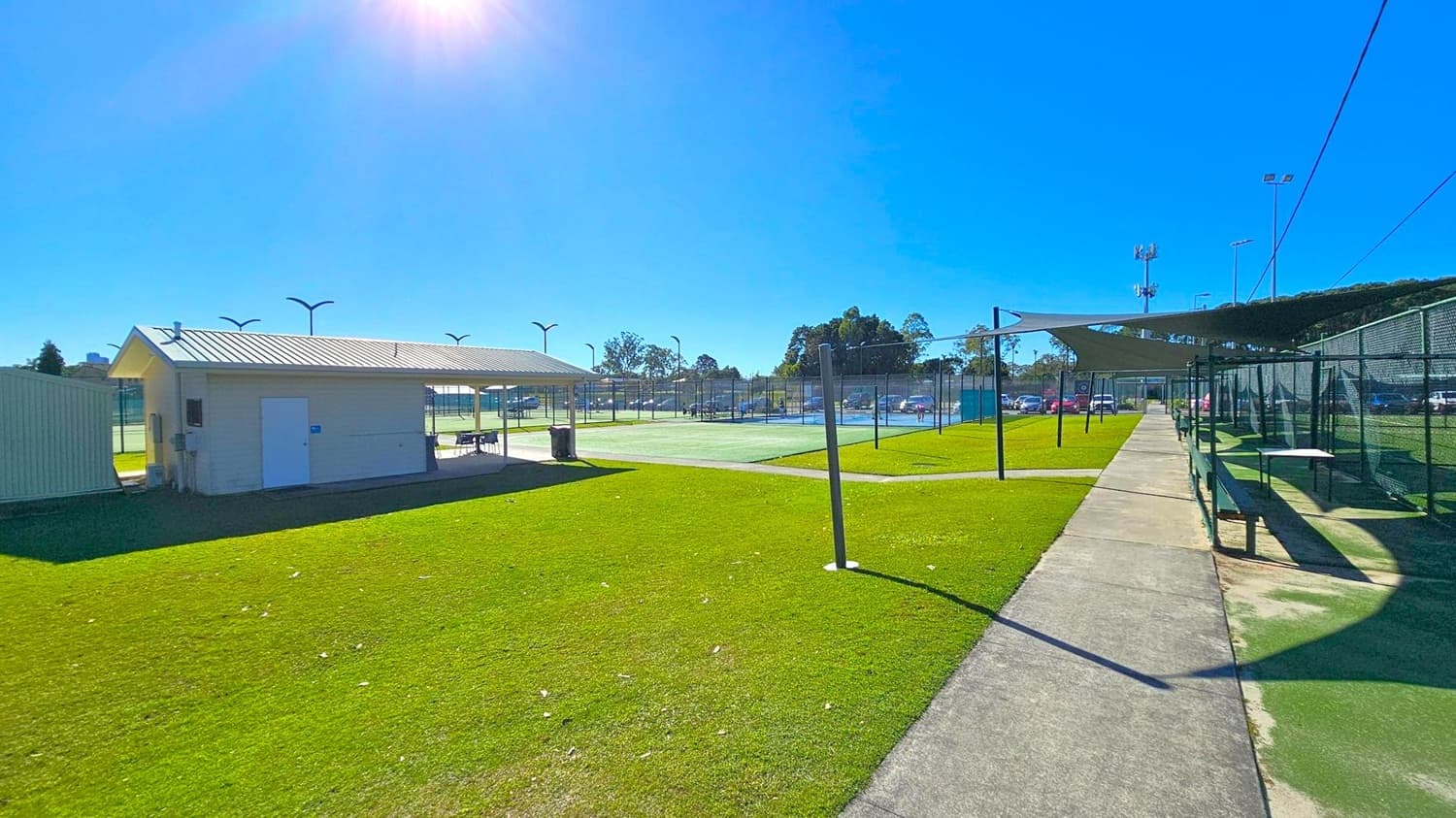 A grassy area with a small building on the left, featuring a metal roof and a covered seating area. In the centre, there are tennis courts enclosed by fencing, with several light poles around. On the right, a paved walkway runs alongside more fenced courts, with shade structures overhead. In the background, trees and parked cars are visible. The sky is clear and sunny.