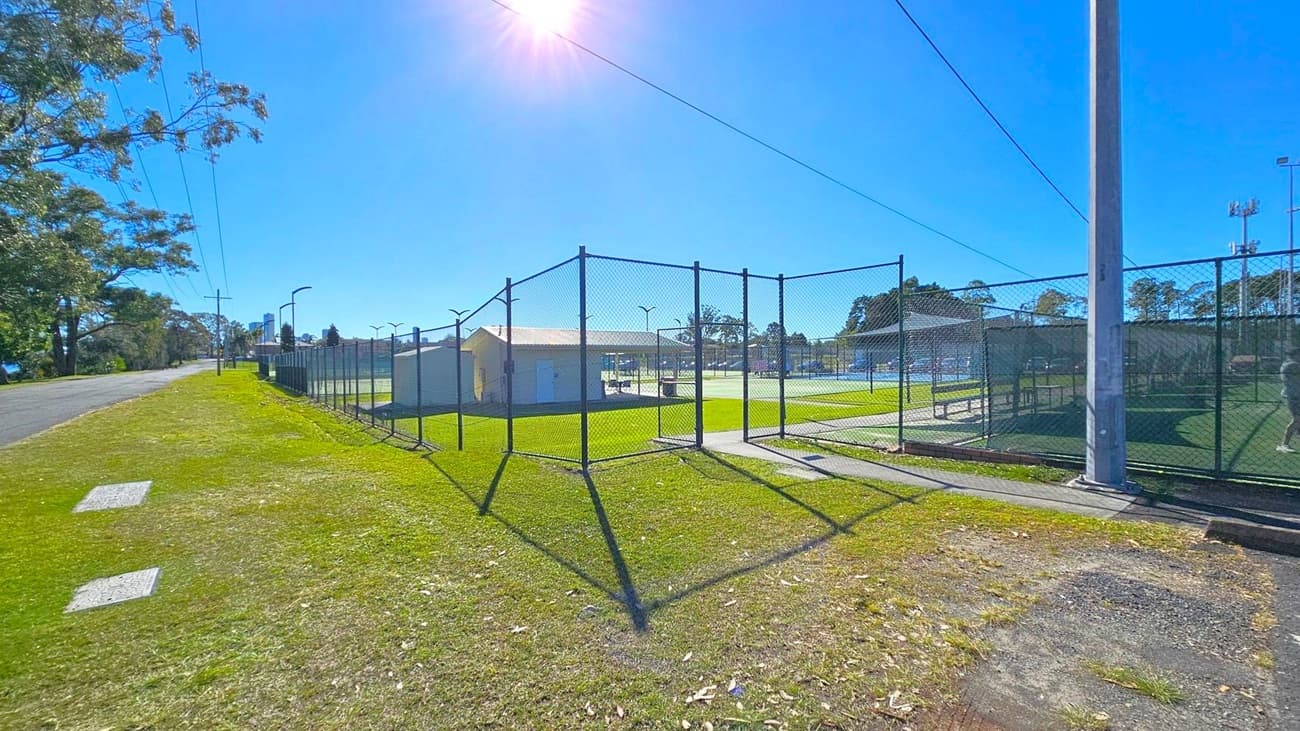 A grassy area with a paved path runs alongside a chain-link fence enclosing tennis courts. The courts are visible through the fence, with a small building in the centre. On the left, a road lined with trees and utility poles extends into the distance. The sky is clear and sunny, casting shadows on the ground.