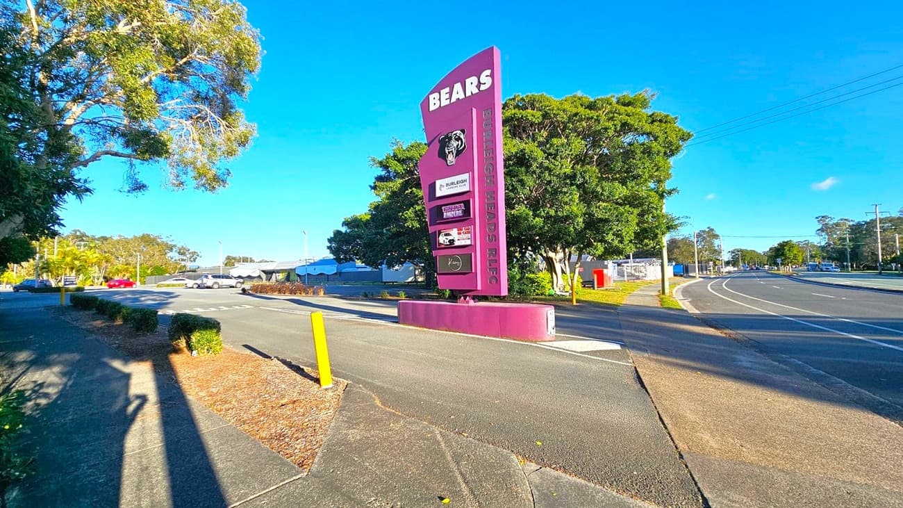 A large purple sign with "BEARS" and "BURLEIGH HEADS RLC" stands prominently on the right, featuring a bear logo and several advertisements. A curved road runs alongside, bordered by a footpath and green grass. On the left, a tree with dense foliage casts shadows on the pavement. In the background, a parking area with a few cars and more trees is visible under a clear blue sky.