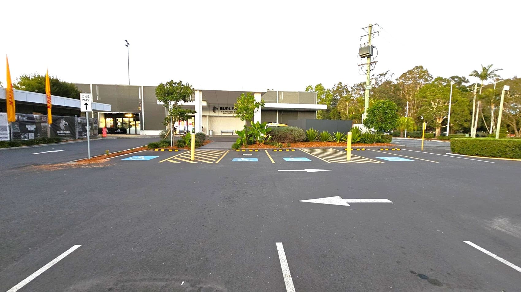 A car park with marked spaces, including disabled parking, is in the foreground. A "One Way" sign and directional arrows are visible on the asphalt. In the background, a building with a sign is flanked by trees and shrubs. Yellow flags are on the left, and a power pole with cables is on the right. The area is surrounded by greenery, including palm trees and hedges.