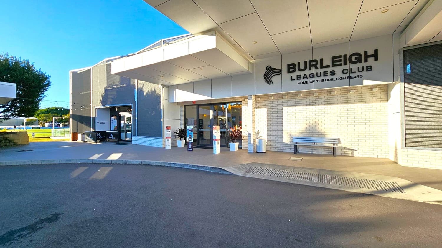 Entrance to a building with a sign reading "Burleigh Leagues Club" and "Home of the Burleigh Bears" on the right. The facade is made of light-coloured bricks. A bench and potted plants are near the entrance. The left side shows a tree and a small outdoor area with string lights. The ground is paved, and the ceiling has a modern, geometric design.