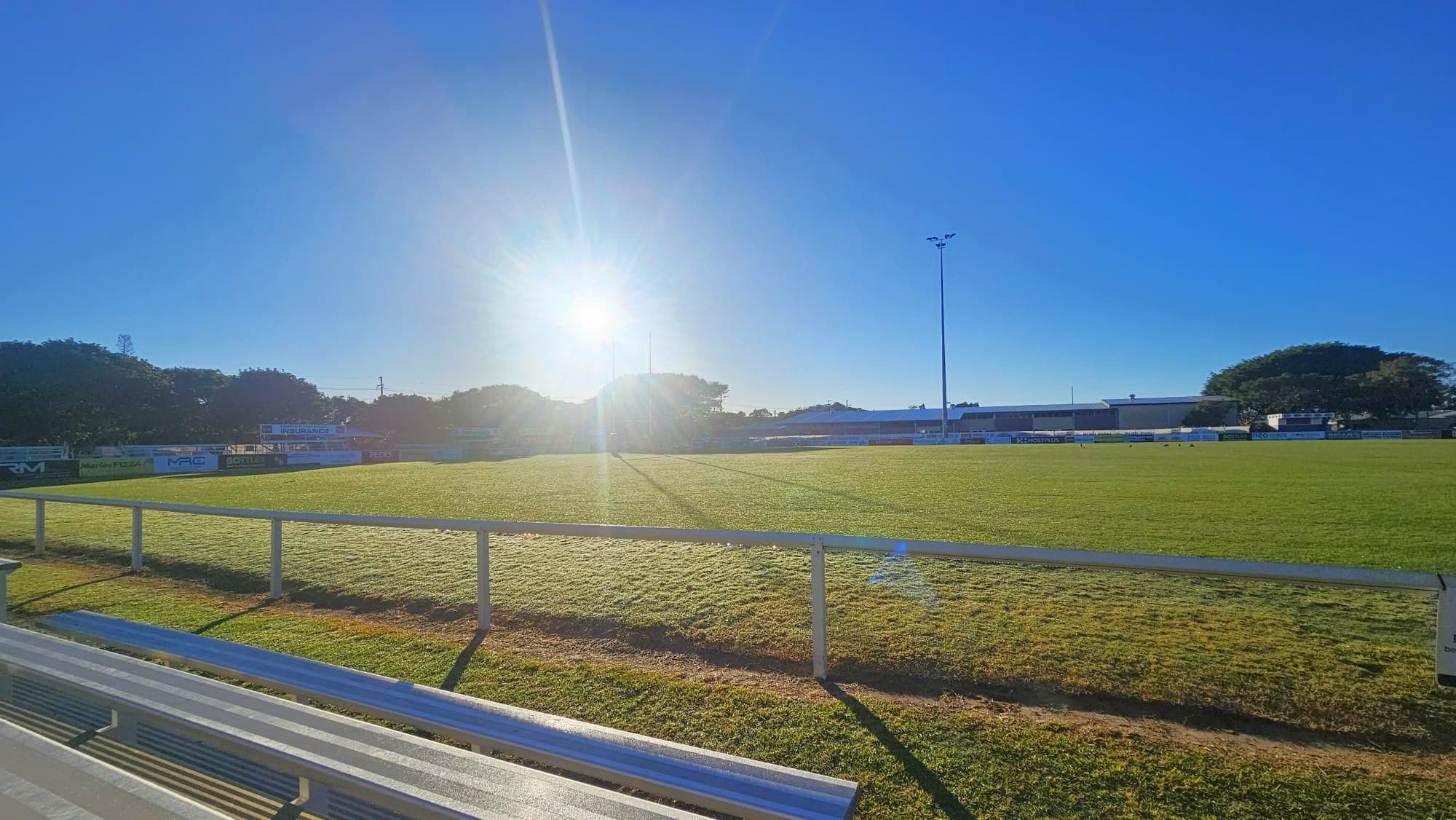A grassy sports field with a low metal railing in the foreground. The sun is low in the sky, casting long shadows across the field. On the left, there are trees and some advertising boards. In the background, a tall floodlight pole is visible, along with more trees and a building. The sky is clear and blue.