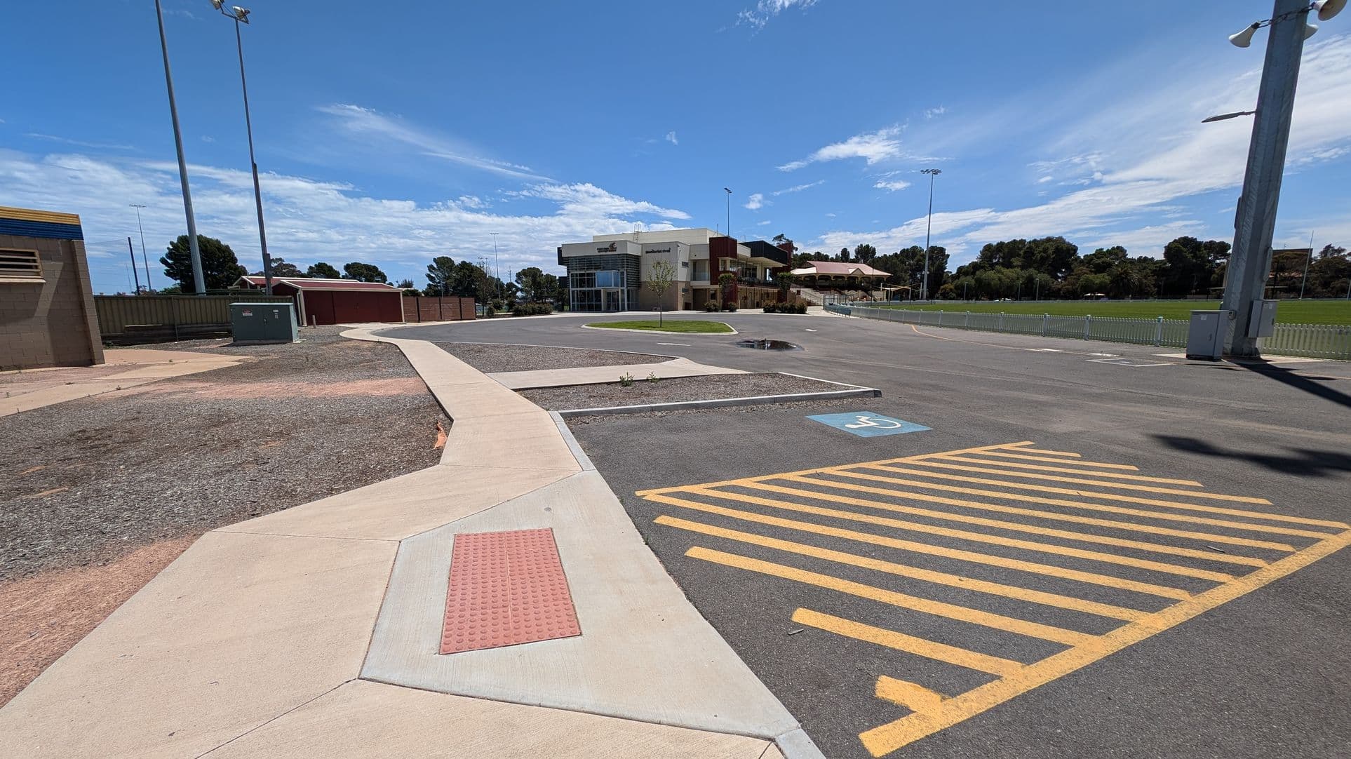 A parking area with two marked disabled parking spaces on the asphalt. Yellow diagonal lines separate the spaces. On the left, a low, beige building with blue trim and a covered walkway. In the background, tall light poles and a fenced area. On the right, a modern building with a curved facade and a small tree on a grassy patch. The ground is mostly paved, with a small puddle visible.