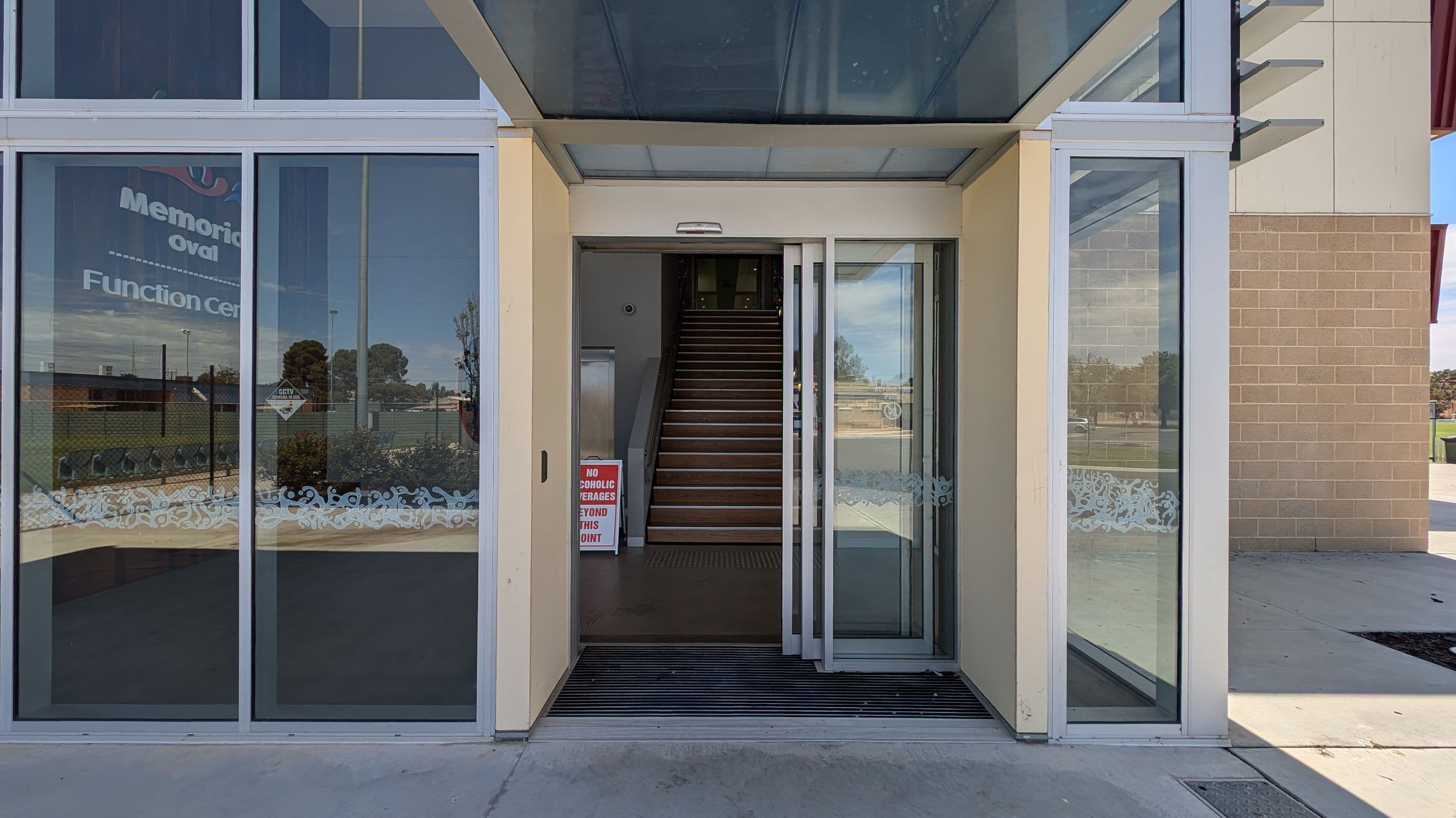 Glass entrance with sliding doors leading to a staircase. On the left, a window displays partial text, "Memorial oval Function Centre," with a view of a grassy area and trees outside. A sign near the entrance reads, "No alcoholic beverages beyond this point." The floor is concrete, and the ceiling above the entrance is glass. The right side shows a brick wall and more outdoor scenery.