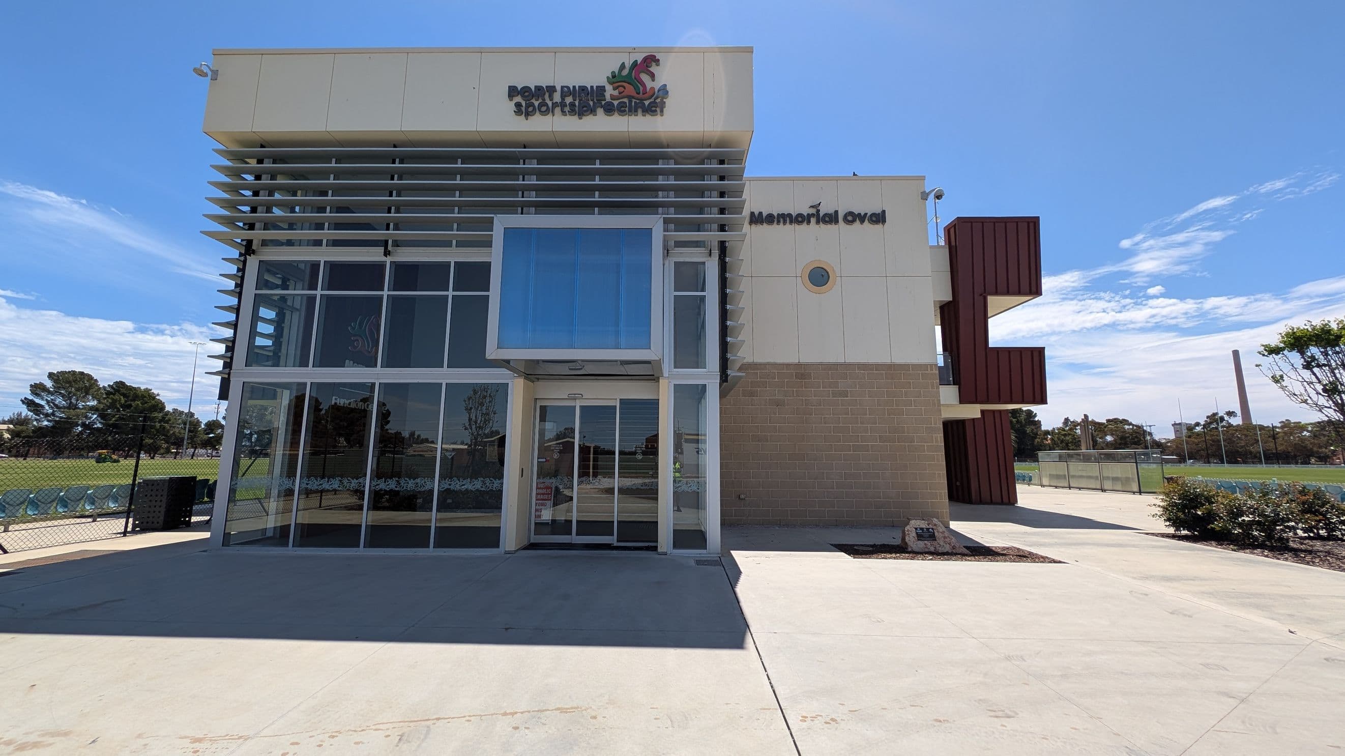 A modern building with a glass facade on the left, featuring horizontal slats and a protruding entrance. The sign reads "Port Pirie Sports Precinct" and "Memorial Oval". The right side has a beige brick wall with a small circular window. In the background, there is a sports field with trees and a tall chimney. The foreground shows a concrete pathway with some shrubs on the right.