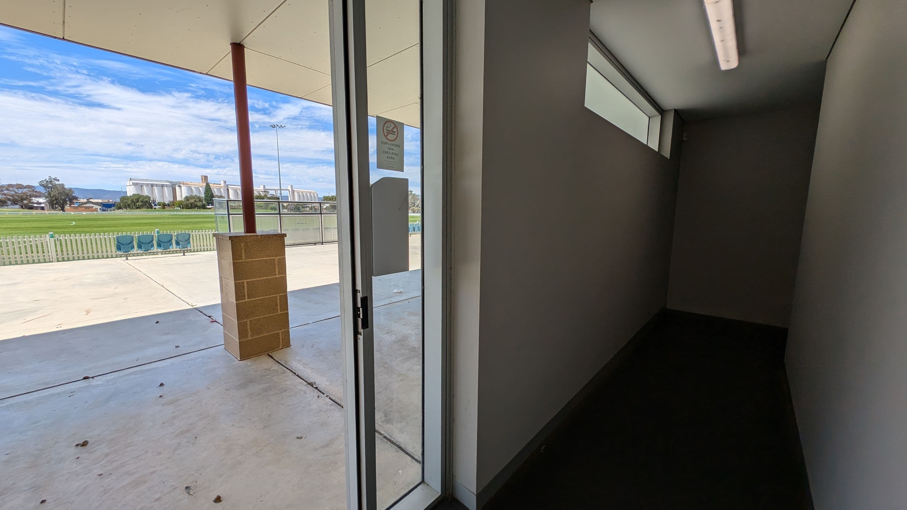 A brick wall with a glass door in the centre, leading to a change room. The door has signs indicating a change room and showers. To the right, a green sign with the number 4 and "Change Room" is mounted on the wall. Below it, an AED (Automated External Defibrillator) is housed in a green box. On the left, a small window is visible above a wooden bench. The floor is concrete.
