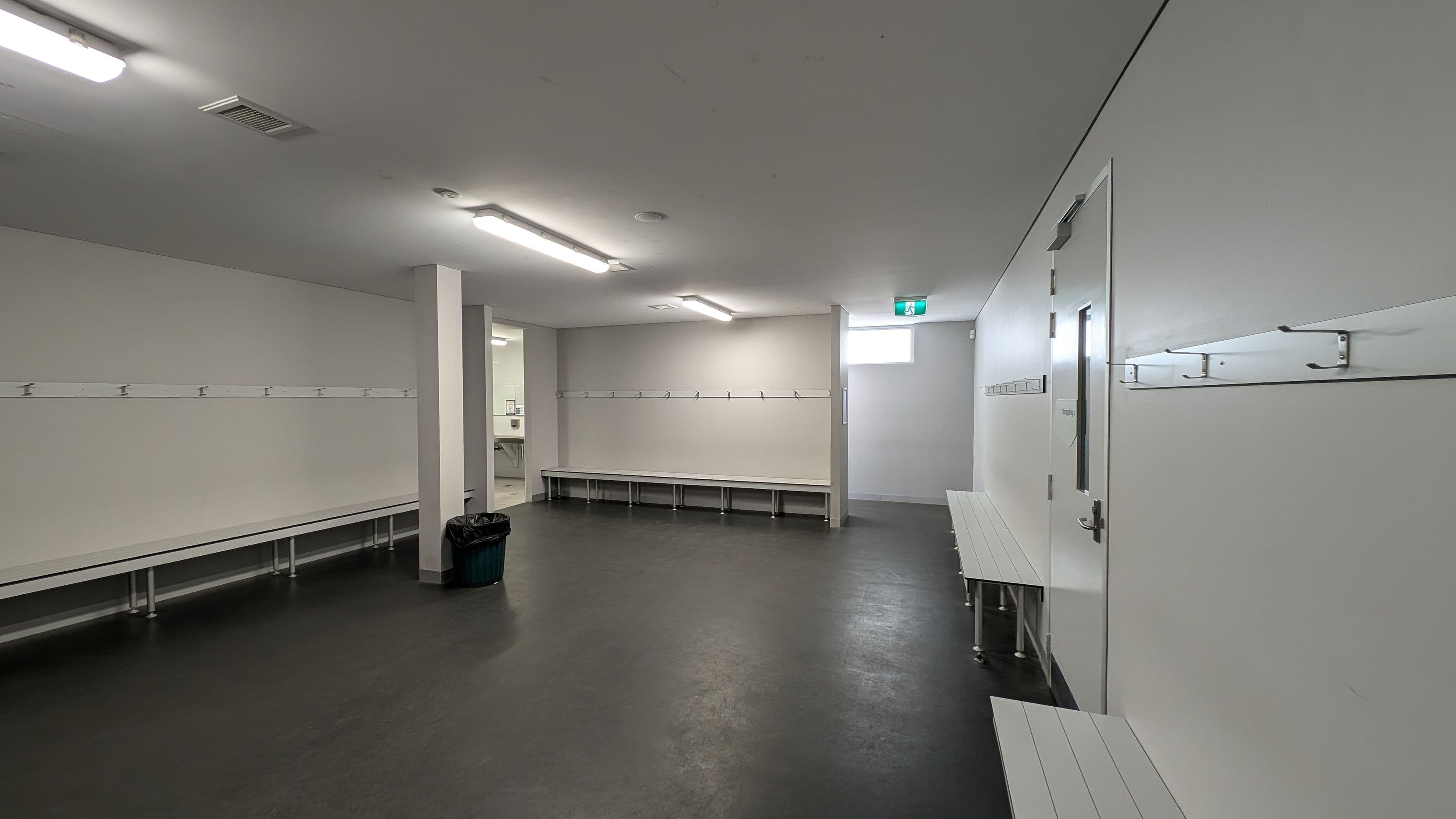 A spacious locker room with grey flooring and white walls. On the left, there are long benches with hooks above them. In the centre, a doorway leads to another room, possibly a bathroom, with visible sinks. On the right, more benches and hooks line the wall, and a door with a small window is present. The ceiling has fluorescent lights, and a small window near the ceiling provides additional light. A green rubbish bin is on the left side of the room.