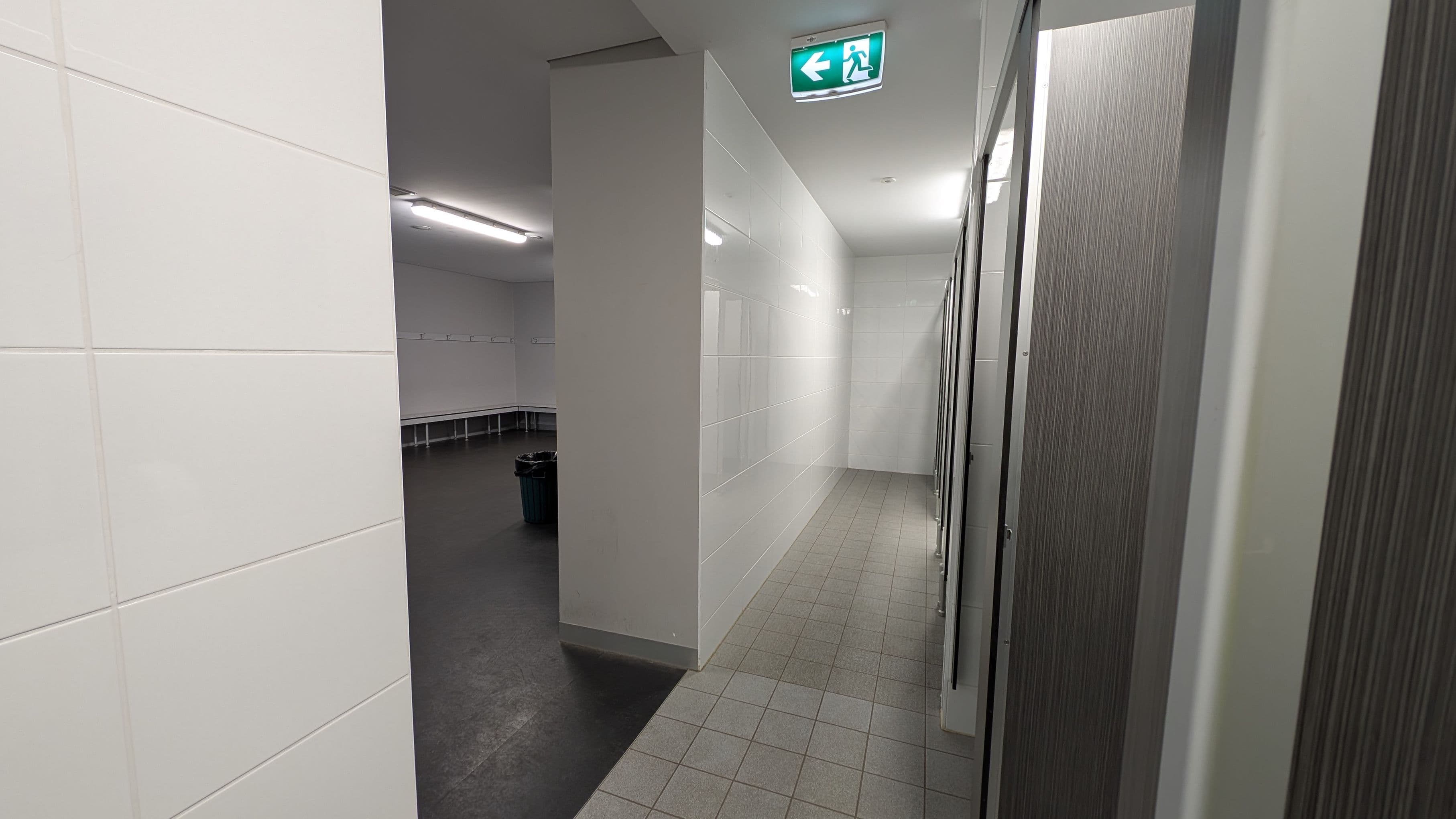 A corridor with white tiled walls and a green-tiled floor leads to a row of cubicles on the right. Above, an illuminated green exit sign with an arrow points left. On the left, an open area with dark flooring features benches along the walls and a rubbish bin. The ceiling is white with fluorescent lighting.