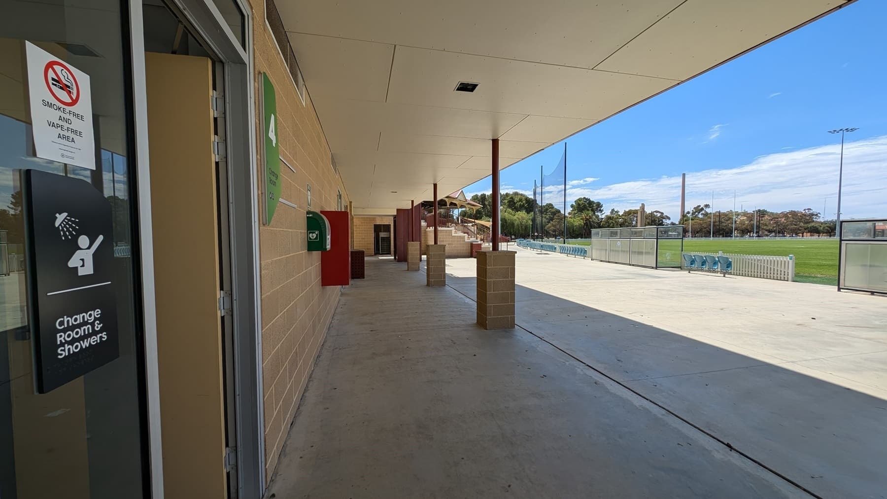 A covered walkway with a beige ceiling and concrete floor. On the left, there's a door with a sign for "Change Room & Showers" and a "Smoke-Free and Vape-Free Area" notice. A green emergency box is mounted on the wall. On the right, an open area with a sports field, seating, and tall light poles. Trees are visible in the background.